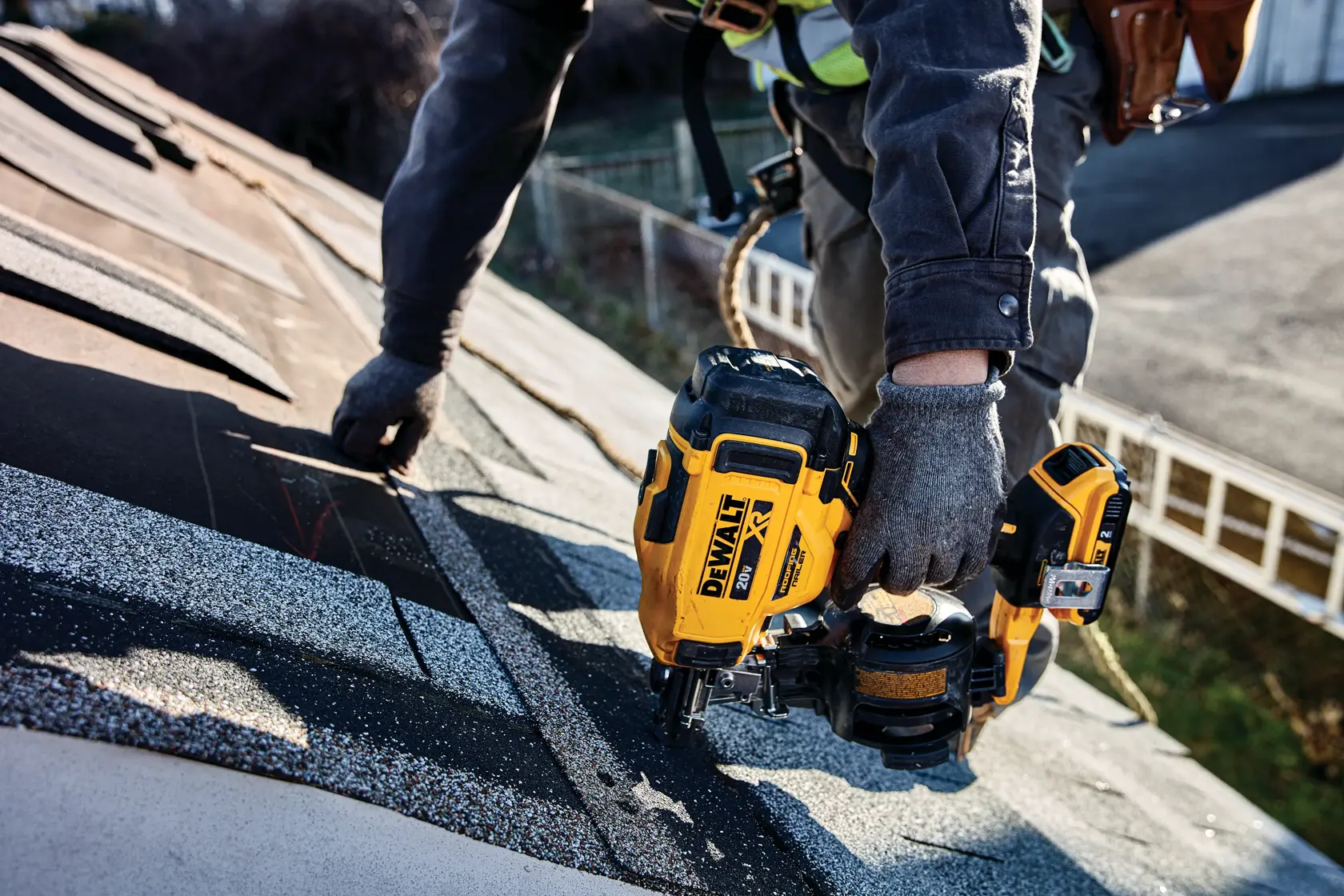 A person wearing work gloves is using a DEWALT cordless roofing nailer on a shingle roof. The tool is yellow and black, and the person is fastening shingles while working on a sloped rooftop.