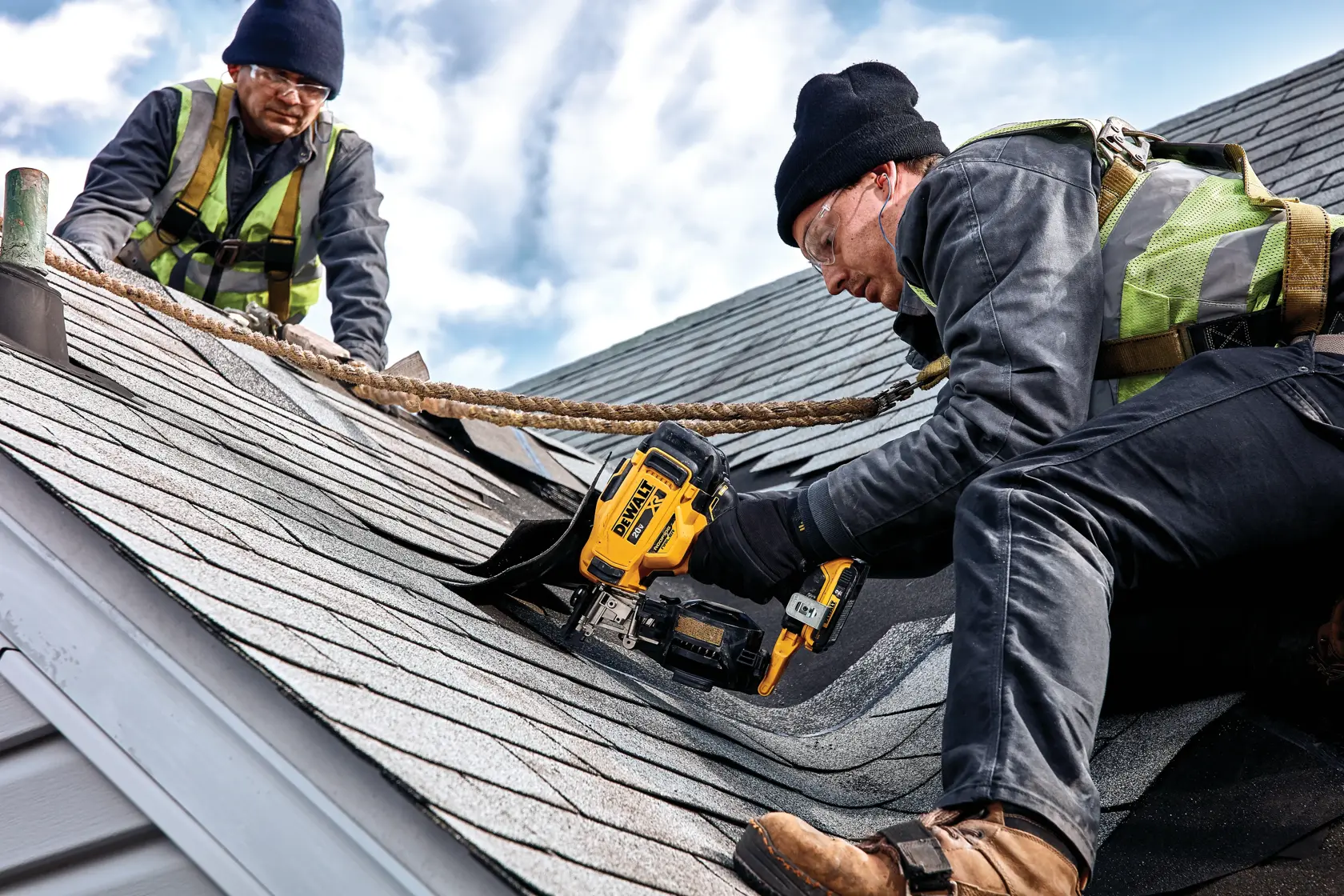 Two workers wearing safety harnesses and vests are installing shingles on a roof. One worker is using a DEWALT nailer tool while the other assists. The scene shows a clear sky and proper safety equipment.