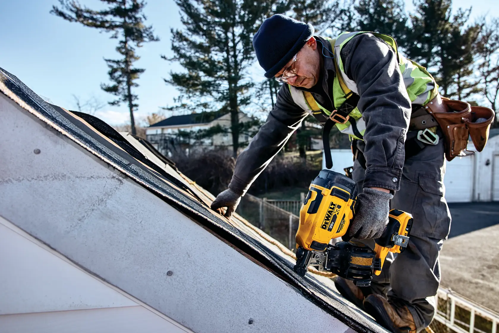 A person wearing work gloves, a safety vest, and a tool belt uses a DEWALT DCN45RND1 nailer to install roofing shingles on a sloped roof. The face of the person is blurred for privacy, and the setting appears to be outdoors with trees and buildings in the background.