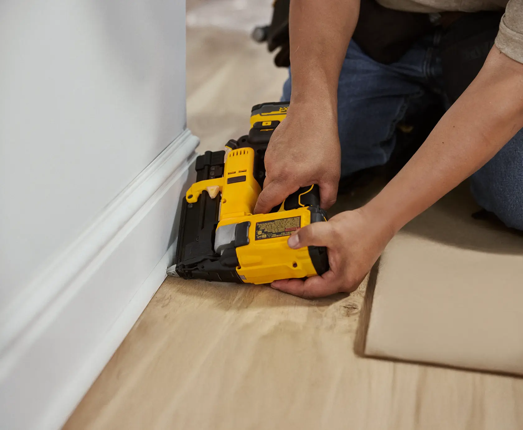 A person kneeling on the floor uses a yellow DEWALT cordless nailer to secure base molding to the wall in a home interior setting.