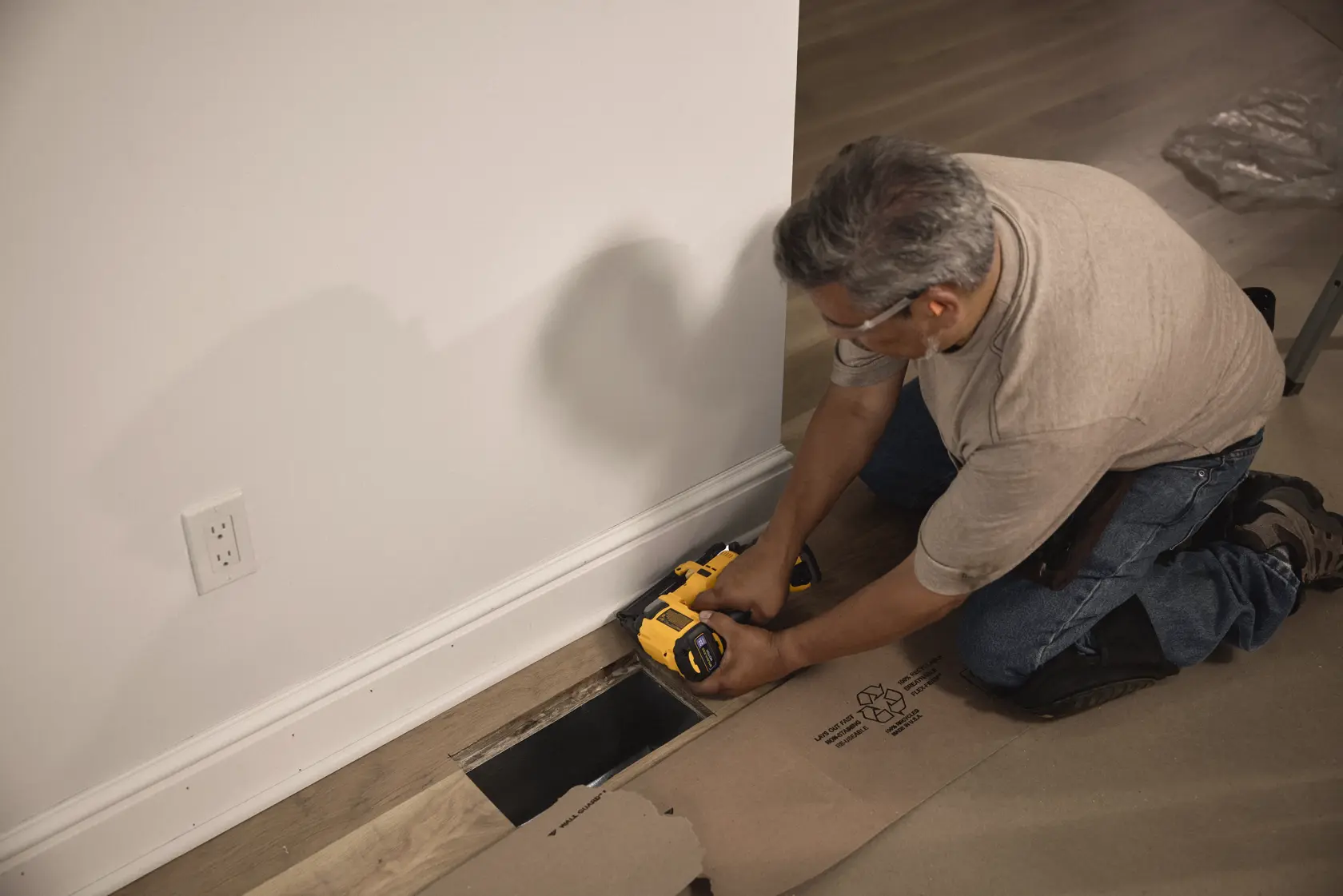 A person using a DEWALT tool to work on floor installation near a wall and a vent opening. The individual is kneeling on the floor, operating the yellow and black tool on a piece of wood.