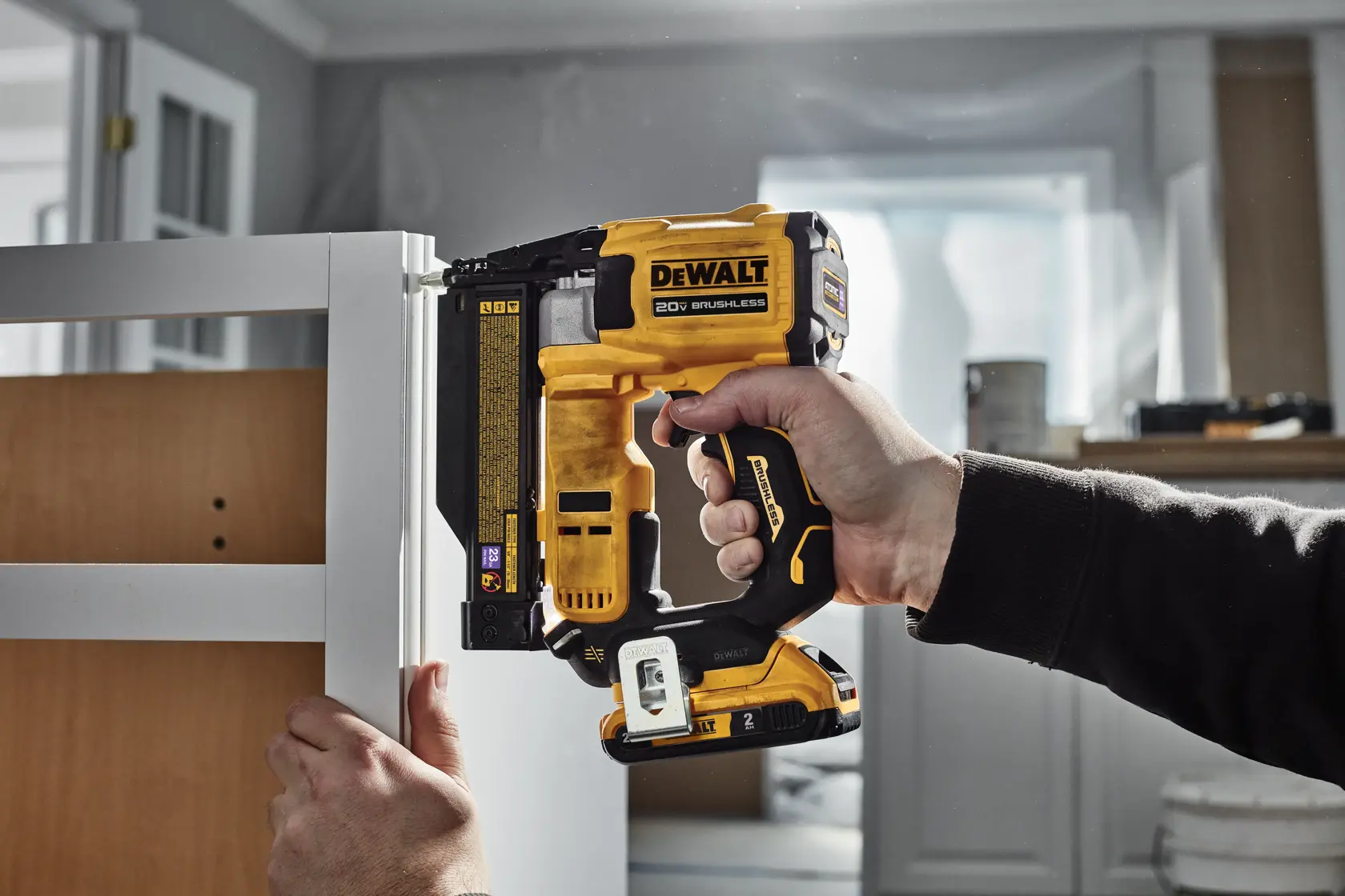A person using a DEWALT 20V cordless brushless nailer to attach a piece of trim to a cabinet or furniture in an indoor workshop setting.