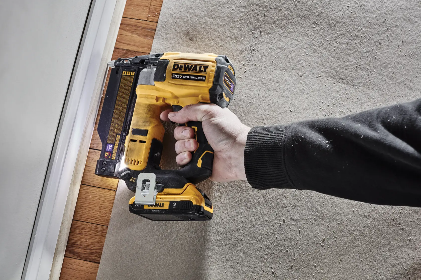 A hand holding a DEWALT 20V brushless cordless nailer, positioned against a baseboard for installation work.