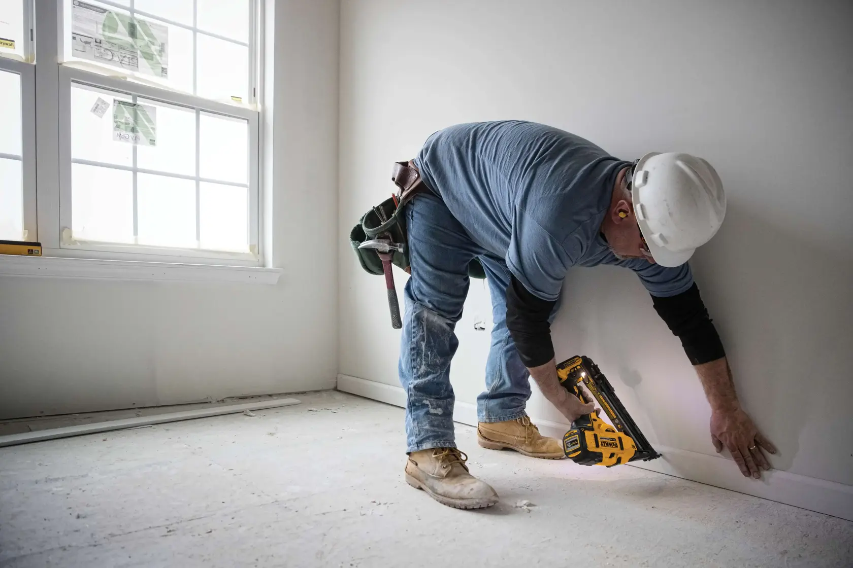 A worker wearing a hard hat and ear protection is using a DEWALT cordless nailer (model DCN650B) to attach trim to the base of a wall in a room under construction. The worker is bending down next to a window and is surrounded by construction tools and materials.