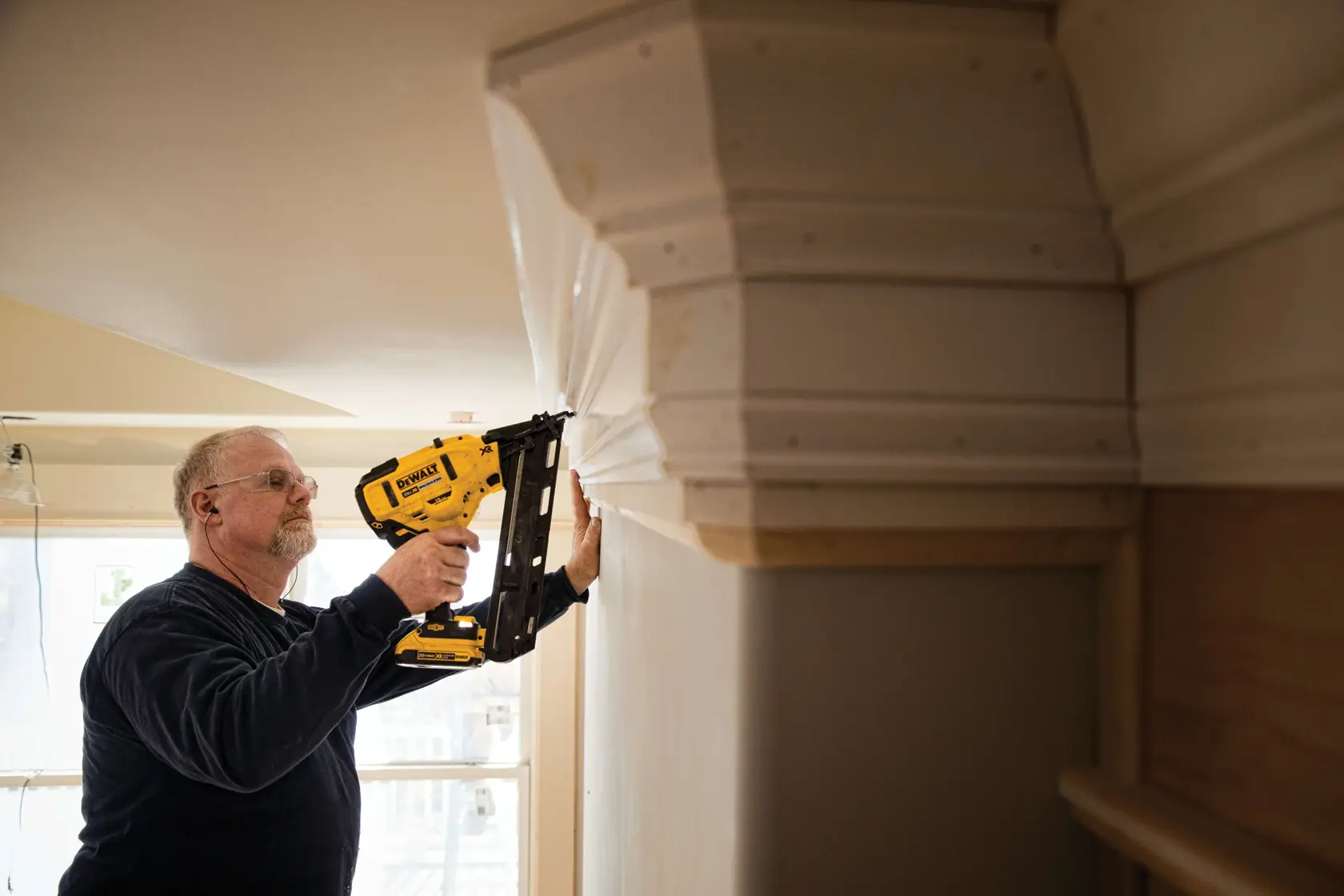 A person using a yellow DEWALT cordless nail gun (model DCN650B) to attach molding to a wall near the ceiling in an indoor construction or renovation setting.