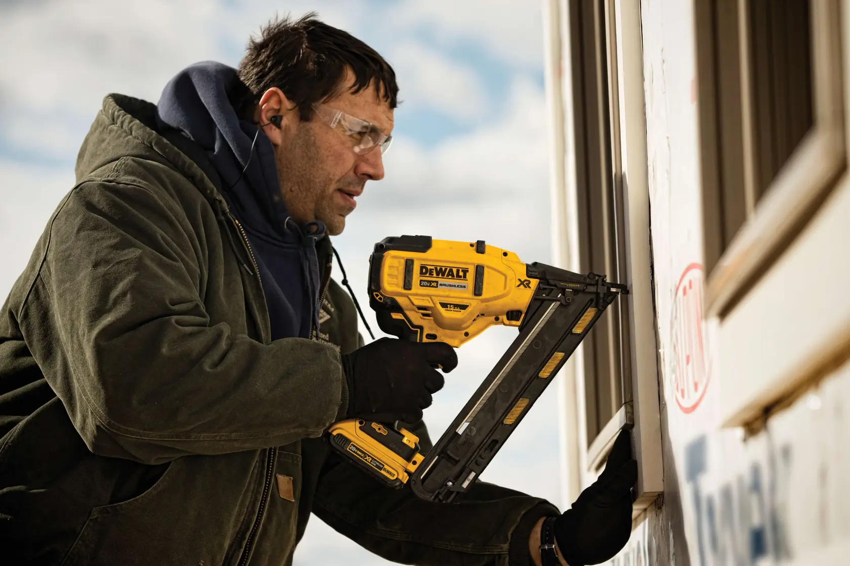 A person wearing a green jacket and gloves uses a DEWALT DCN650B cordless nailer to secure trim on the exterior of a building.
