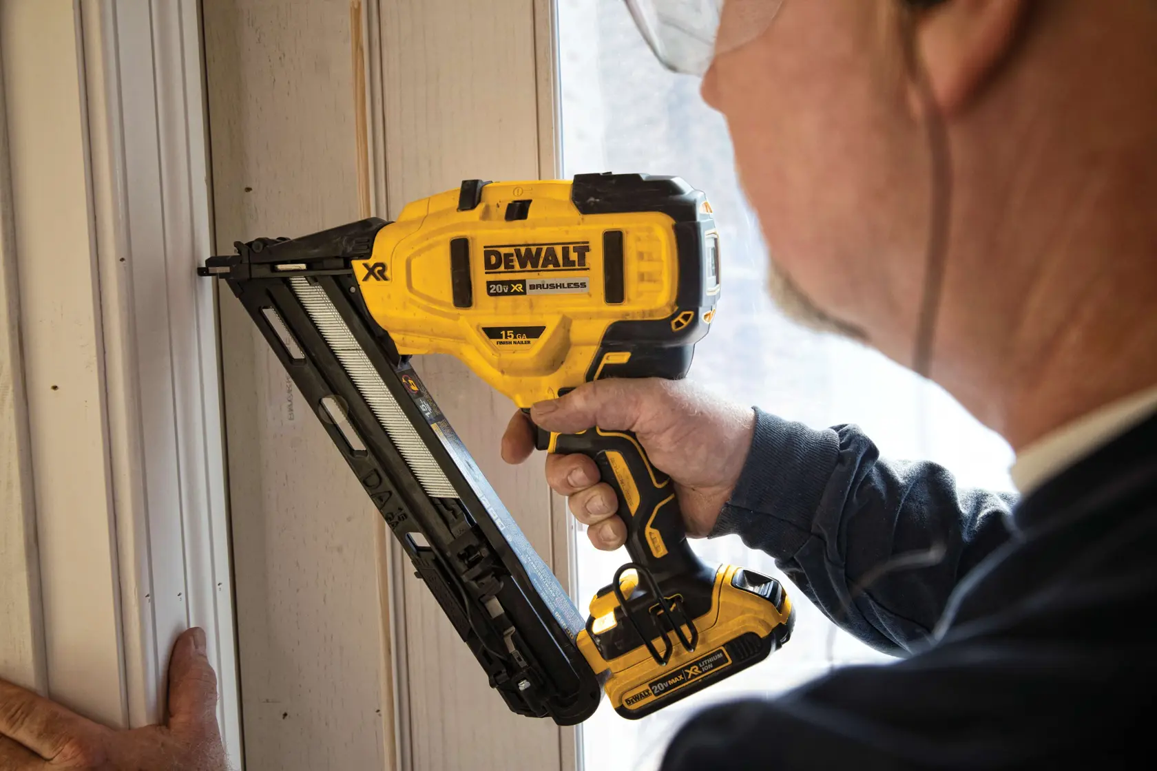 A person using a yellow DEWALT cordless nail gun to install trim on a wooden wall.