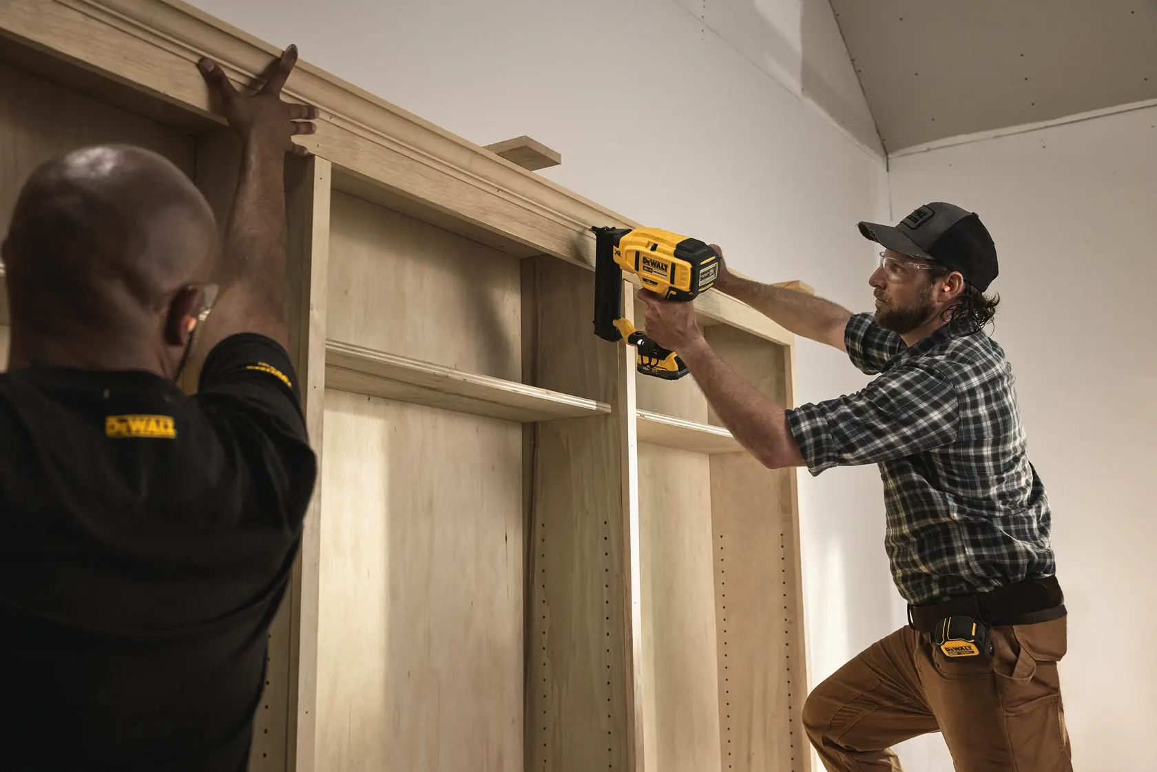 Two people are installing wooden trim on a cabinet using a DEWALT cordless nail gun. One person is holding the wood in place while the other is operating the nail gun.
