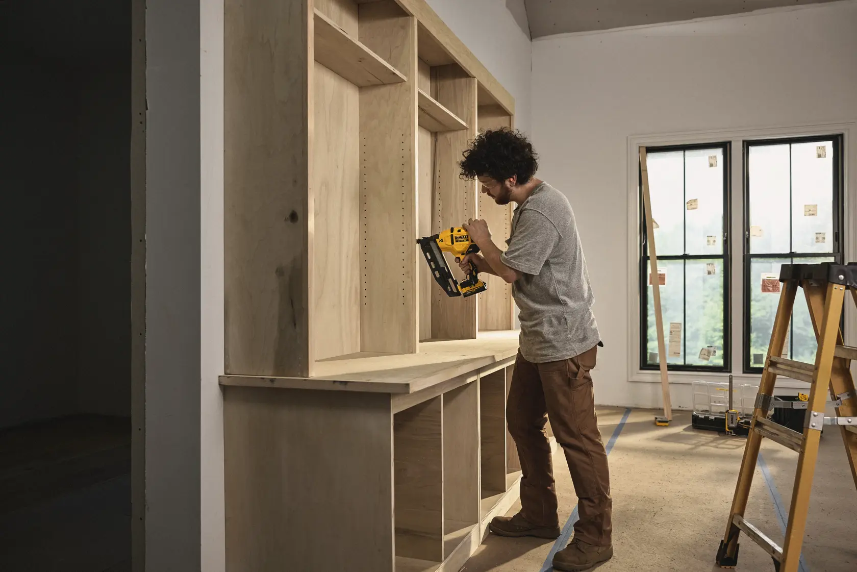 A person is using a DEWALT cordless nailer to assemble wooden shelving in a room under construction. The person is standing near a ladder and a window, wearing work clothes.