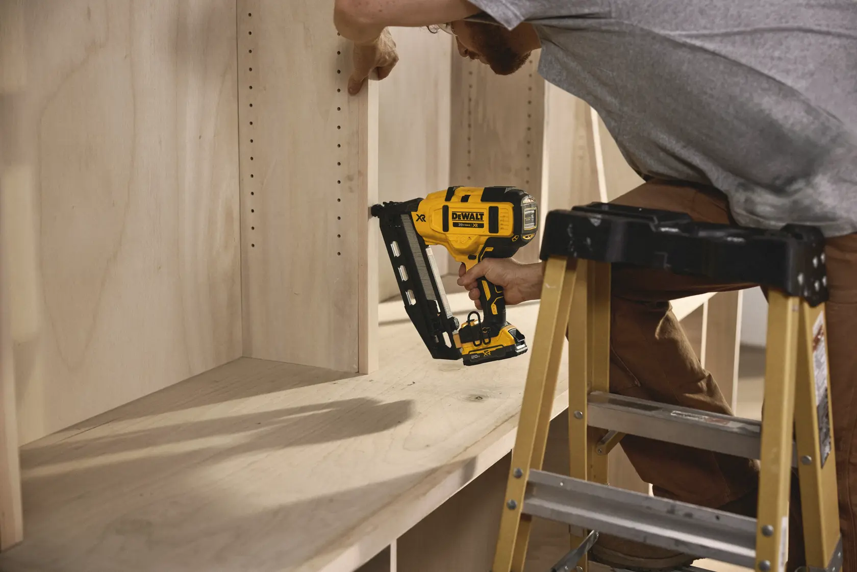 A person uses a DEWALT cordless nail gun (model DCN660B) to fasten a wooden panel inside a shelving unit. The person is standing on a step ladder and working carefully with the tool.