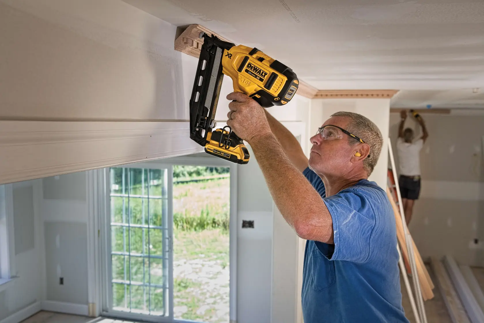 A person using a DEWALT DCN660D1 cordless nailer to install molding on the ceiling of a room under construction. Another person is working in the background.