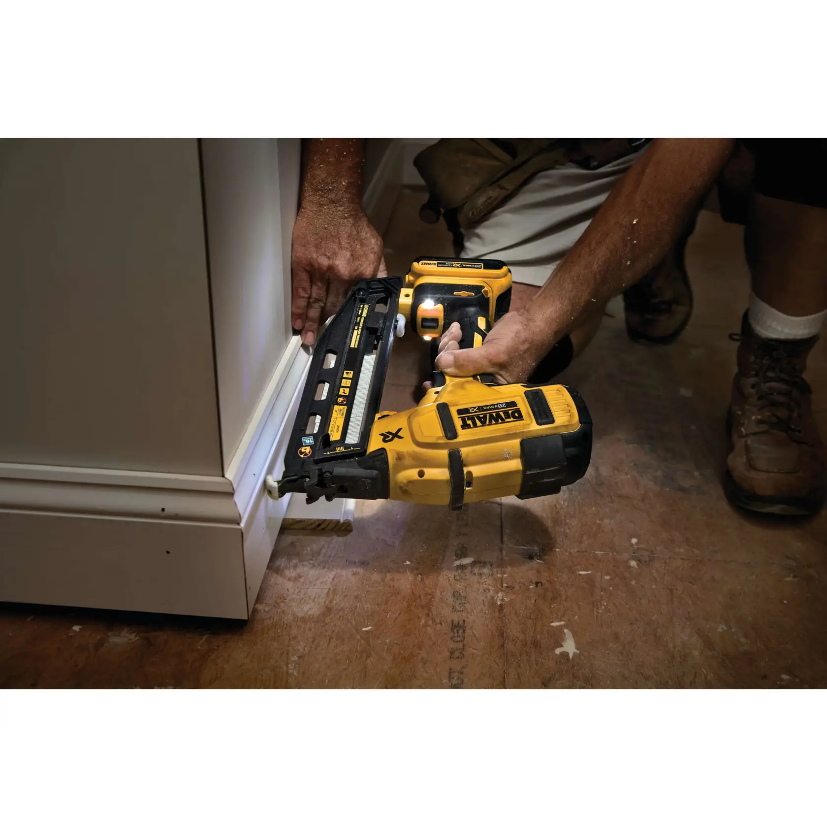 A person is using a yellow DEWALT cordless nail gun (DCN660D1) to attach trim or molding to the bottom edge of a cabinet on a wooden floor.