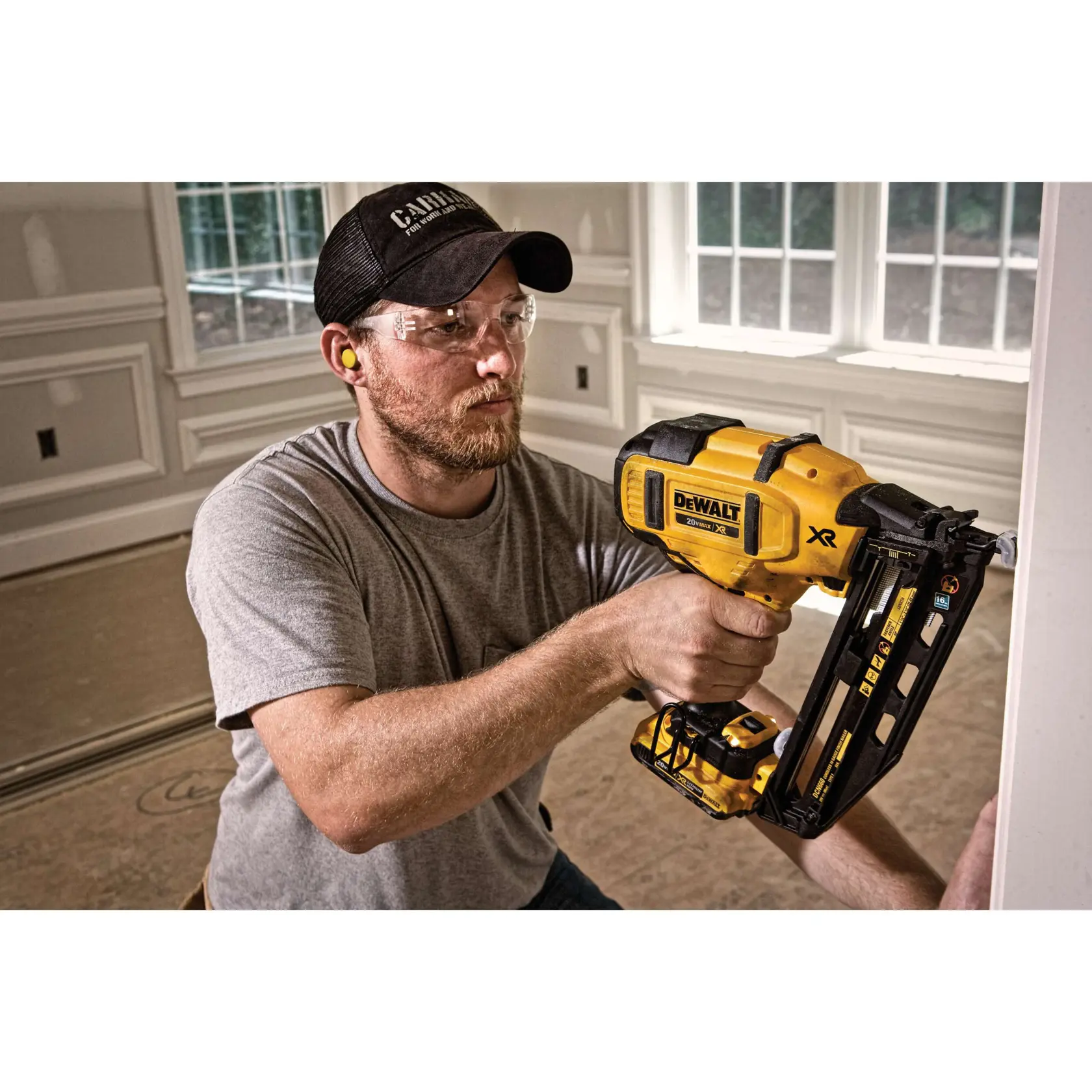 A person with a blurred face is using a DEWALT XR cordless nailer to install trim in a room with large windows and white molding. The setting appears to be a construction or renovation site.