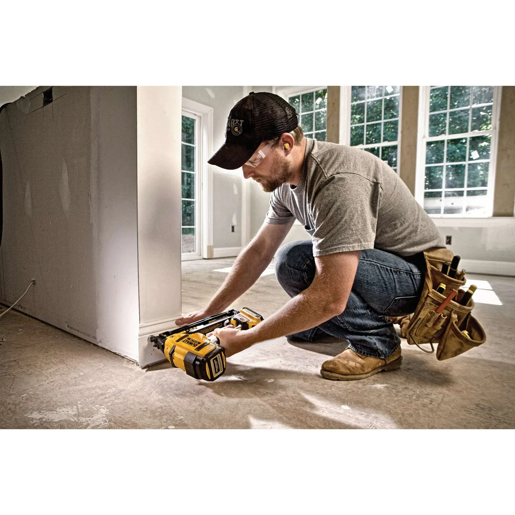 A person kneeling on the floor is using a DEWALT cordless nailer to attach trim to the base of a wall in a bright, unfinished room. The person is wearing work boots, jeans, a gray t-shirt, a black cap, and a tool belt holding various tools. The face of the person is blurred.