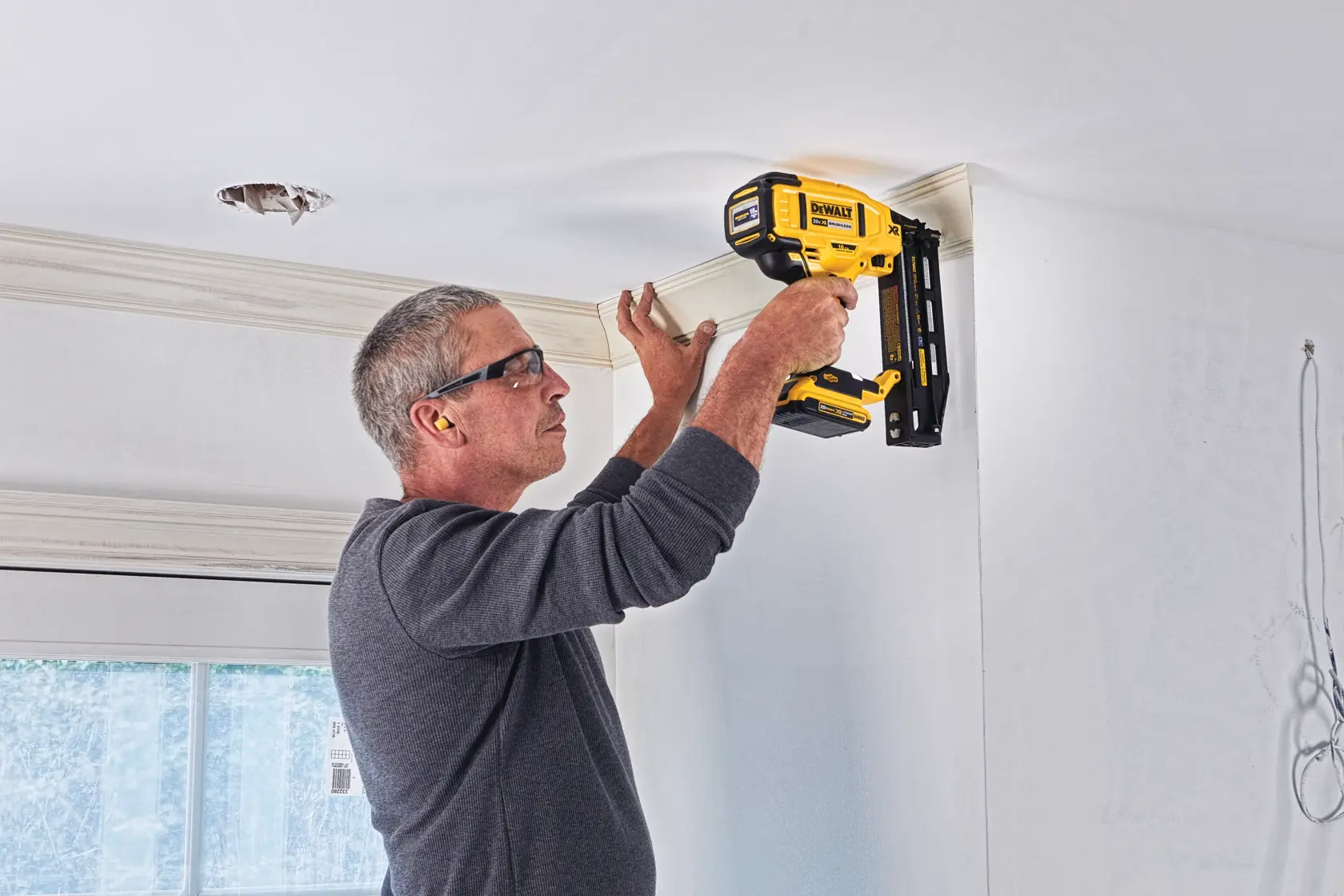 A person using a yellow DEWALT cordless nailer (model DCN662D1) to install crown molding on a white ceiling in a room.