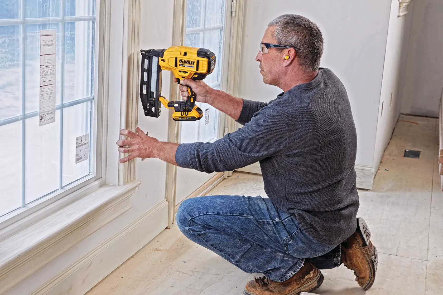 A person is kneeling while using a DEWALT cordless nailer to install trim around a window. The individual's face is blurred, and they are wearing ear protection. The scene is indoors, in a room with light-colored walls and large windows.