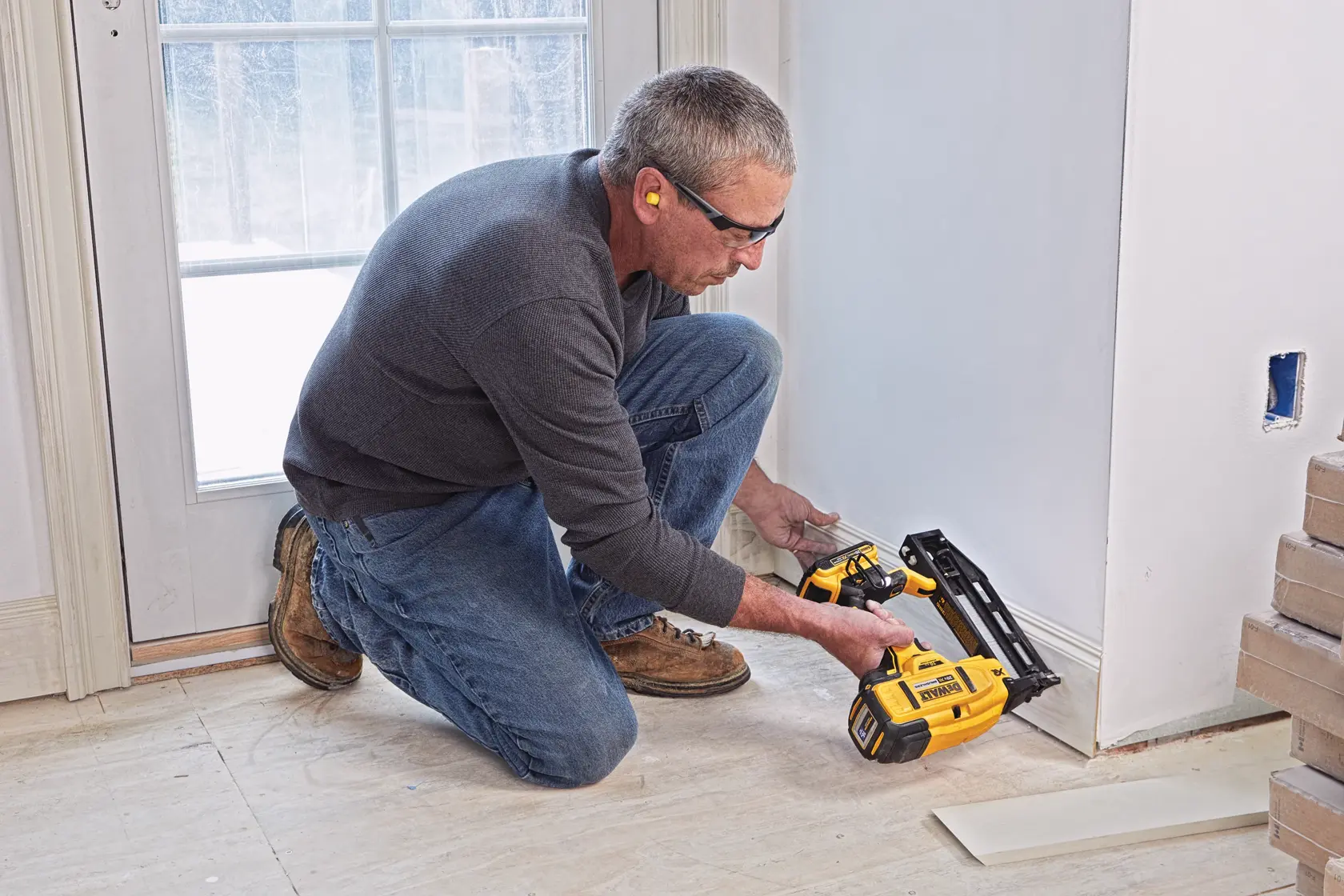 A person kneels on the floor using a DEWALT cordless nailer to install baseboard trim near a wall and doorway. The person's face is blurred for privacy.
