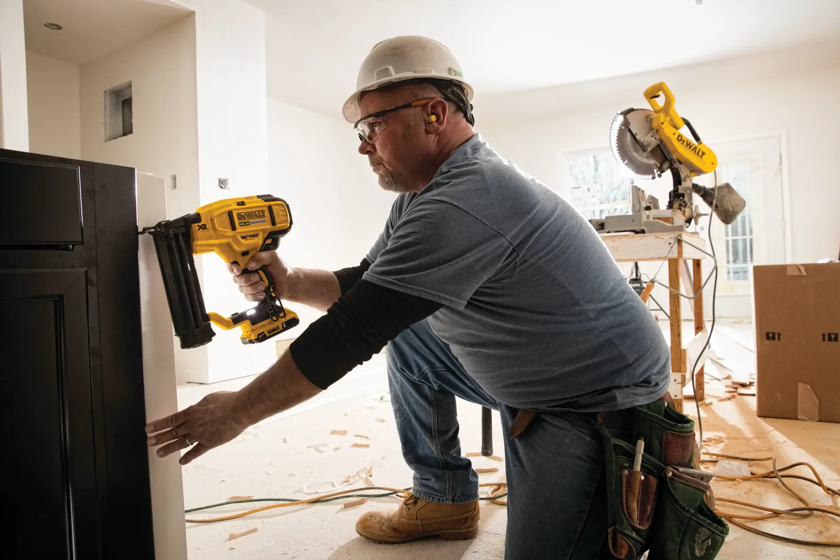 A person wearing a hard hat and ear protection is using a yellow DEWALT nail gun (model DCN680B) to attach a cabinet or panel indoors. The person is kneeling on the floor in a construction or renovation setting, with wood shavings and power tools visible in the background.