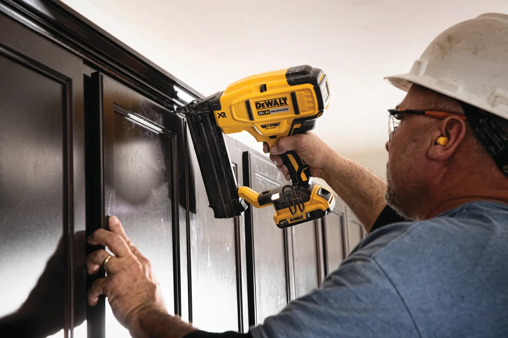 A person using a DEWALT DCN680D1 cordless nailer to install molding on dark wooden cabinets. The person is wearing a hard hat, glasses, ear protection, and a blue shirt.