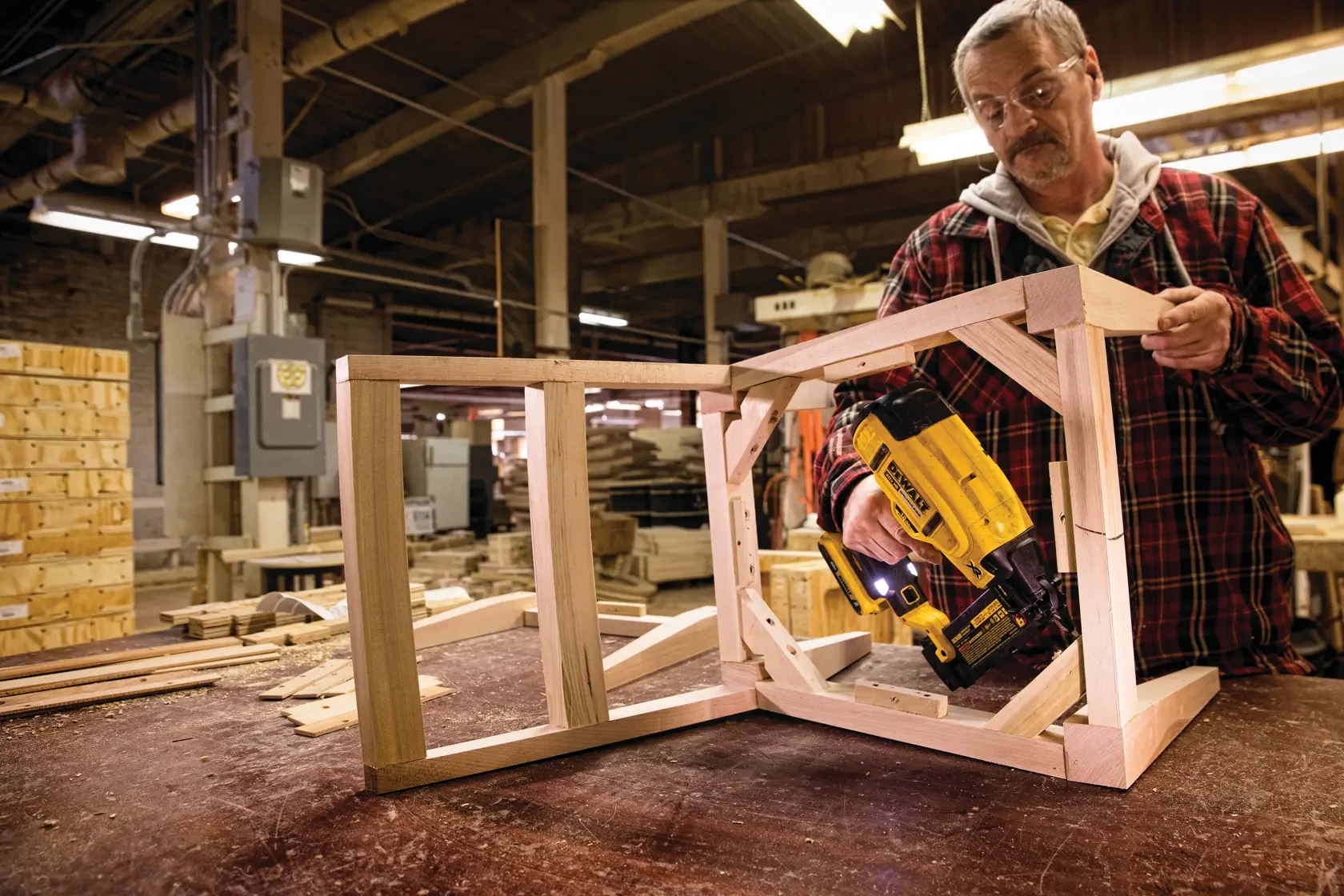 A person using a DEWALT DCN680D1 nail gun to construct a wooden frame in a woodworking shop. The workspace features wood materials and machinery in the background. The person's face is blurred.