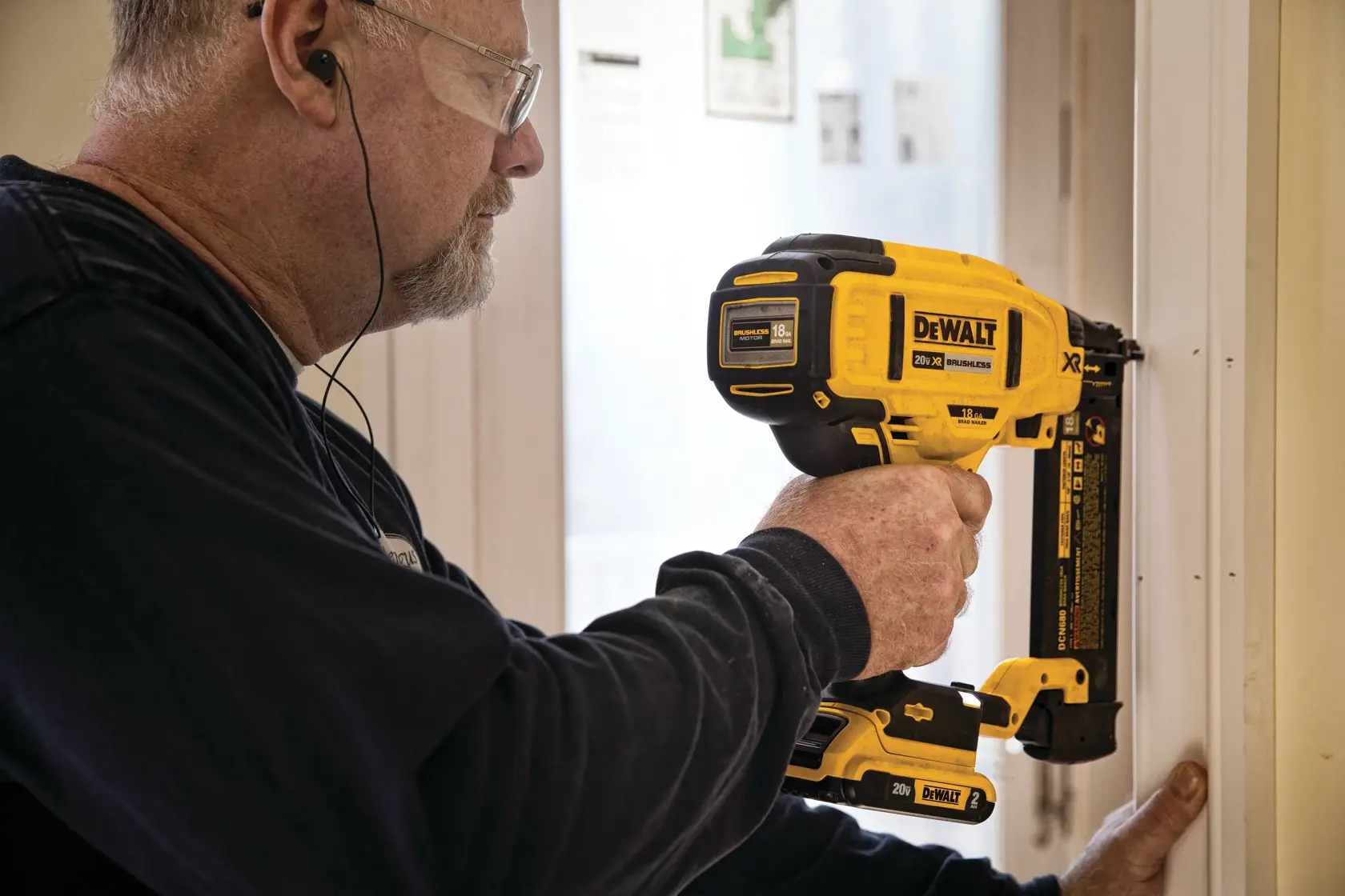 A person using a yellow DEWALT DCN680D1 cordless nailer to attach trim to a door frame. The person's face is blurred for privacy.
