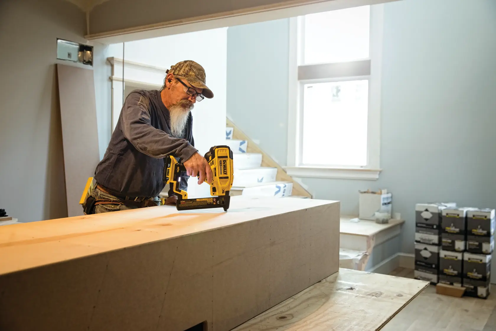 A person is using a DEWALT DCN681B nailer to assemble wood in an indoor workspace. Boxes of materials are visible in the background, and the person is wearing work attire and a cap.