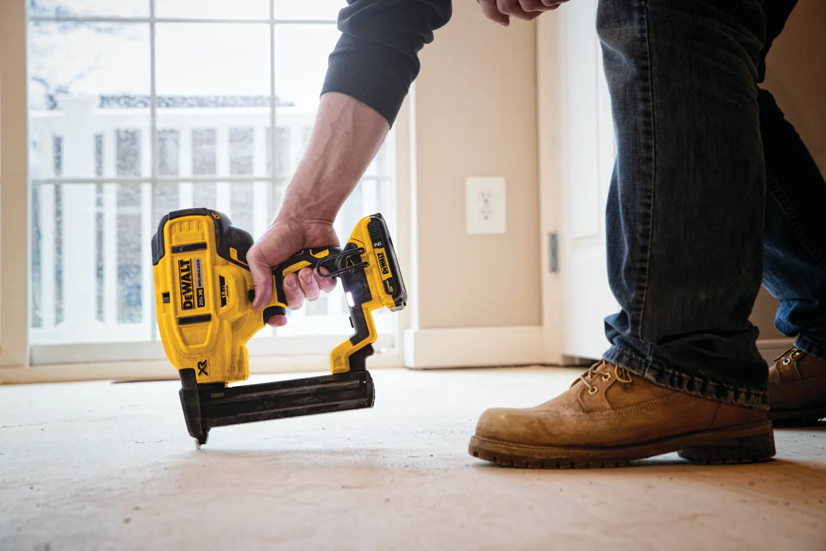 A person using a yellow DEWALT DCN681D1 cordless nailer on a floor indoors, with natural light coming from a nearby window.