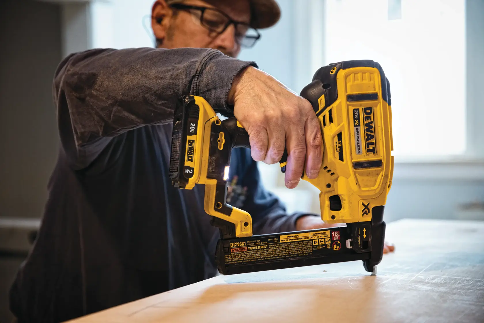 A person using a yellow DEWALT DCN681D1 cordless nailer to fasten wood in an indoor workspace. The tool is being operated with one hand and is positioned against a flat wooden surface.