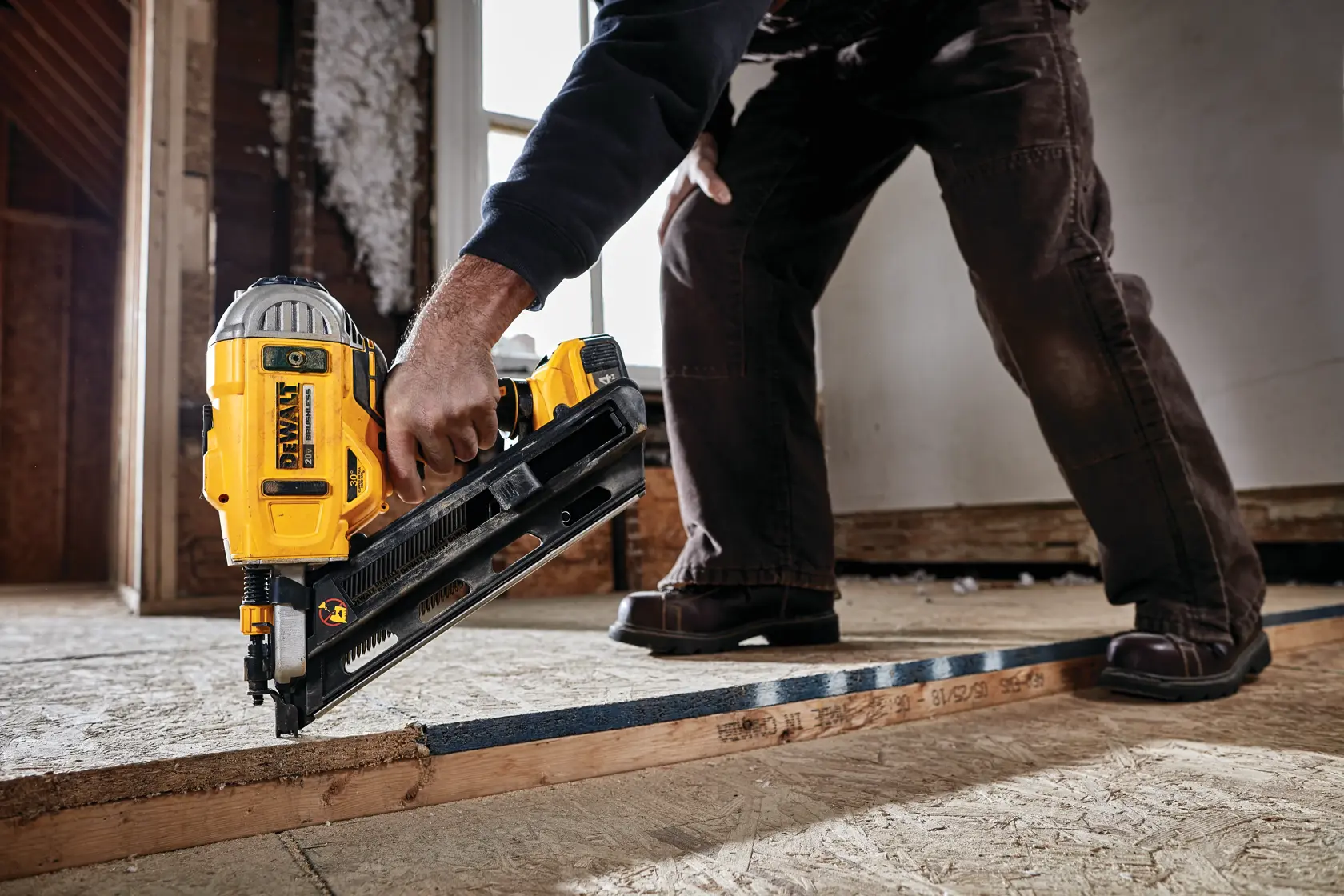 A person using a DEWALT DCN692B cordless nail gun to fasten a wooden beam during construction work indoors. The tool is yellow and black, and the person is wearing dark pants and brown shoes.