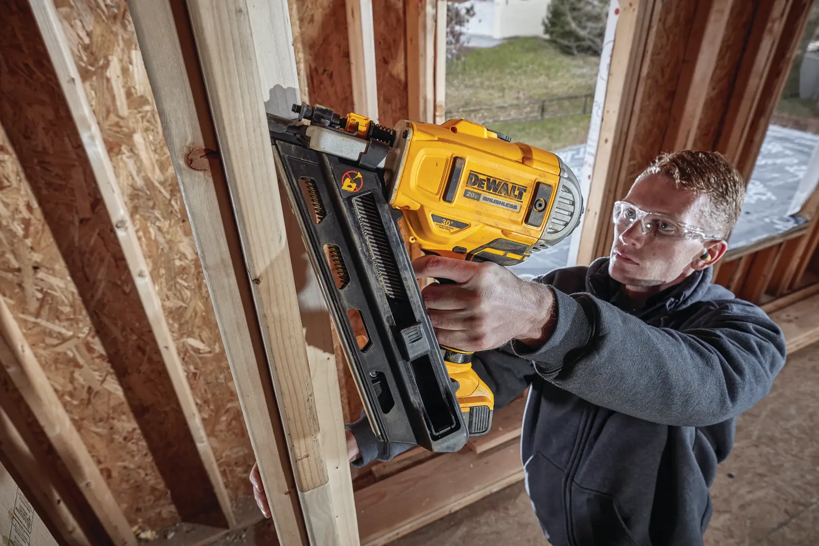 A person using a DEWALT DCN692B cordless nail gun to fasten wooden studs inside a building under construction. The individual’s face is blurred for privacy. The scene is indoors and shows unfinished wooden framing.