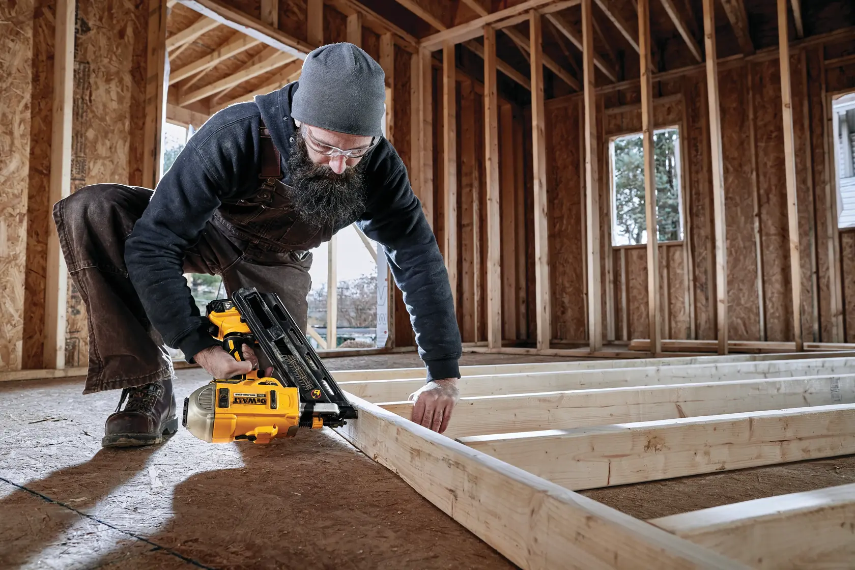 A person wearing work clothes and a beanie is using a DEWALT DCN692M1 cordless nail gun to secure wooden framing in the interior of a building under construction. The person's face is blurred, and the wooden structure is only partially finished.