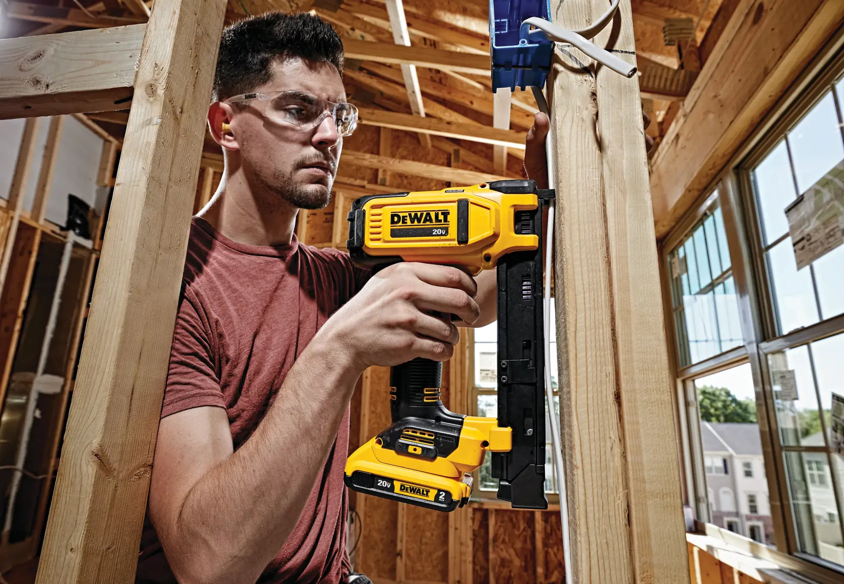 A person using a DEWALT 20V cordless cable stapler (model DCN701D1) in a wooden framed construction area, with their face blurred for privacy.