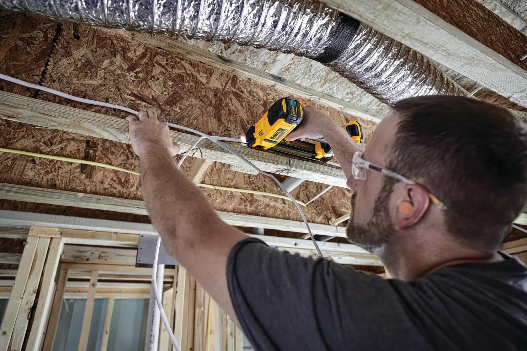 A person using a yellow DEWALT DCN701D1 tool to secure electrical cables onto wooden beams during construction in a building with exposed framing and ductwork. The face of the person is blurred.