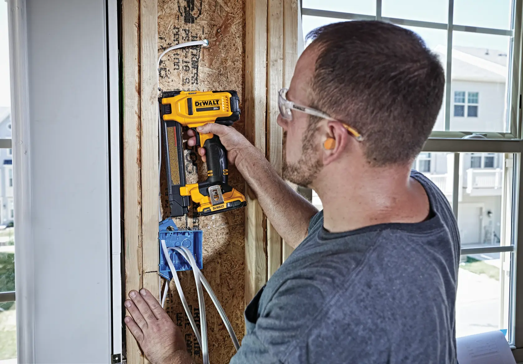 A person using a DEWALT DCN701D1 cordless tool to install cable staples in a wooden frame near a window, with electrical wiring visible and a partially finished wall.