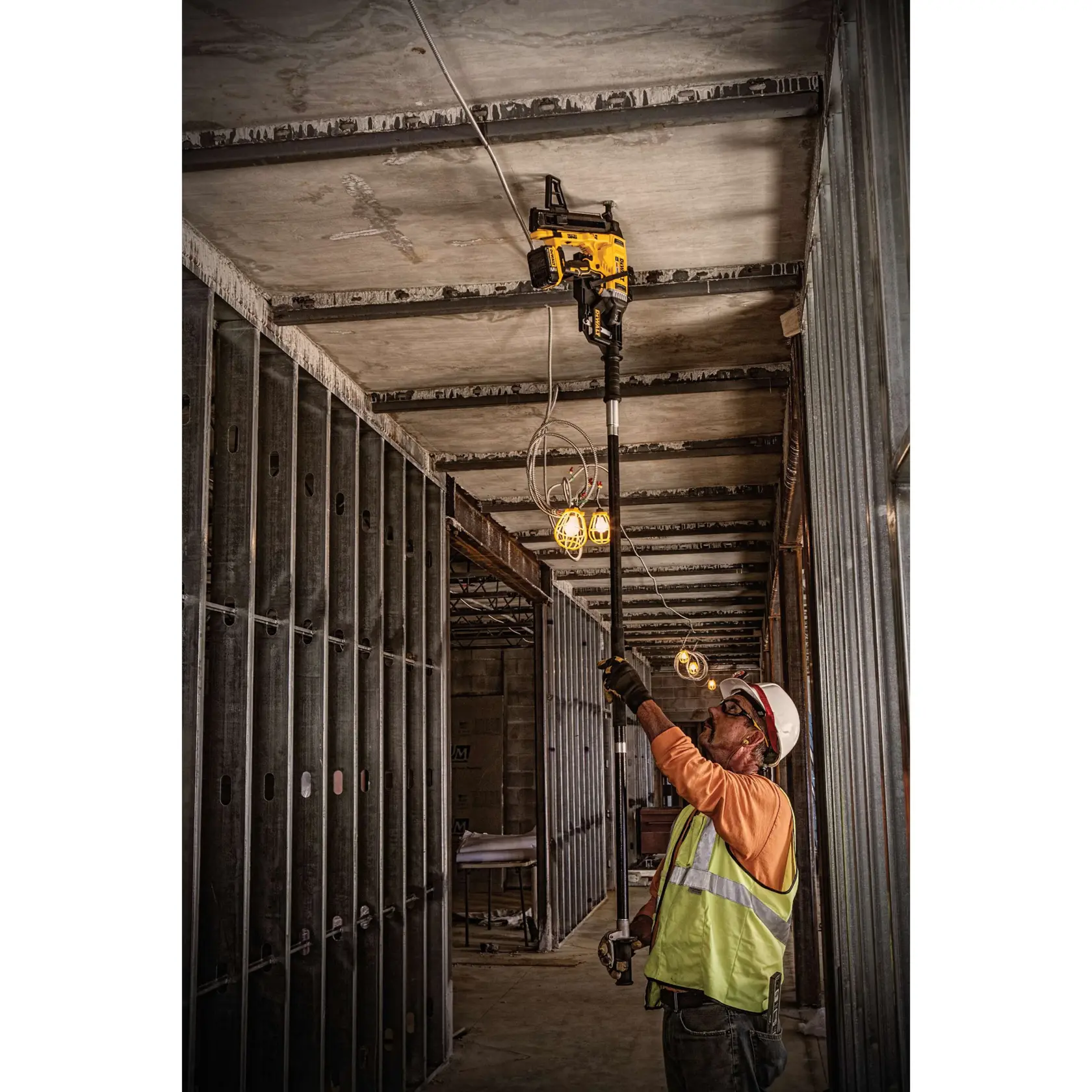 A construction worker wearing a safety vest and helmet uses a DEWALT nailer tool with an extension pole to fasten materials to the overhead concrete ceiling in an unfinished building interior.