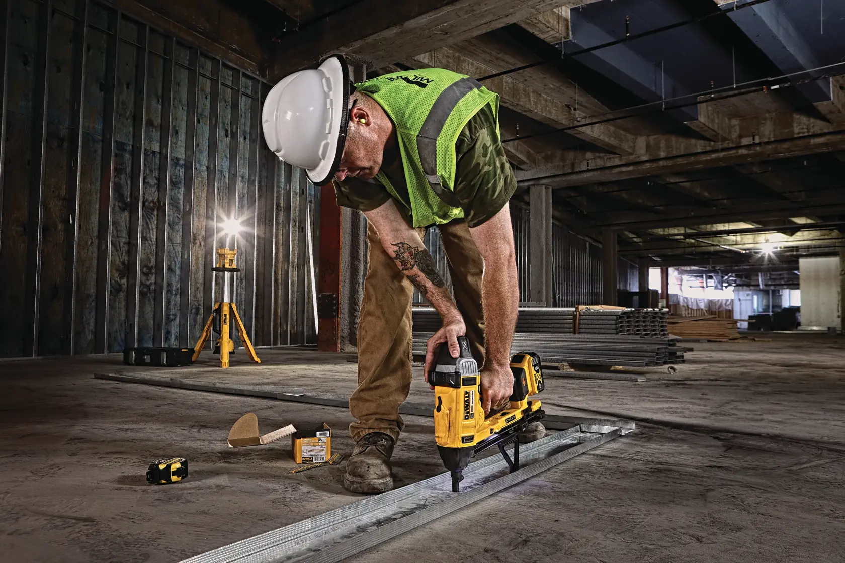A construction worker wearing a hard hat and safety vest uses a yellow DEWALT tool to fasten metal tracks to the floor in an unfinished building.