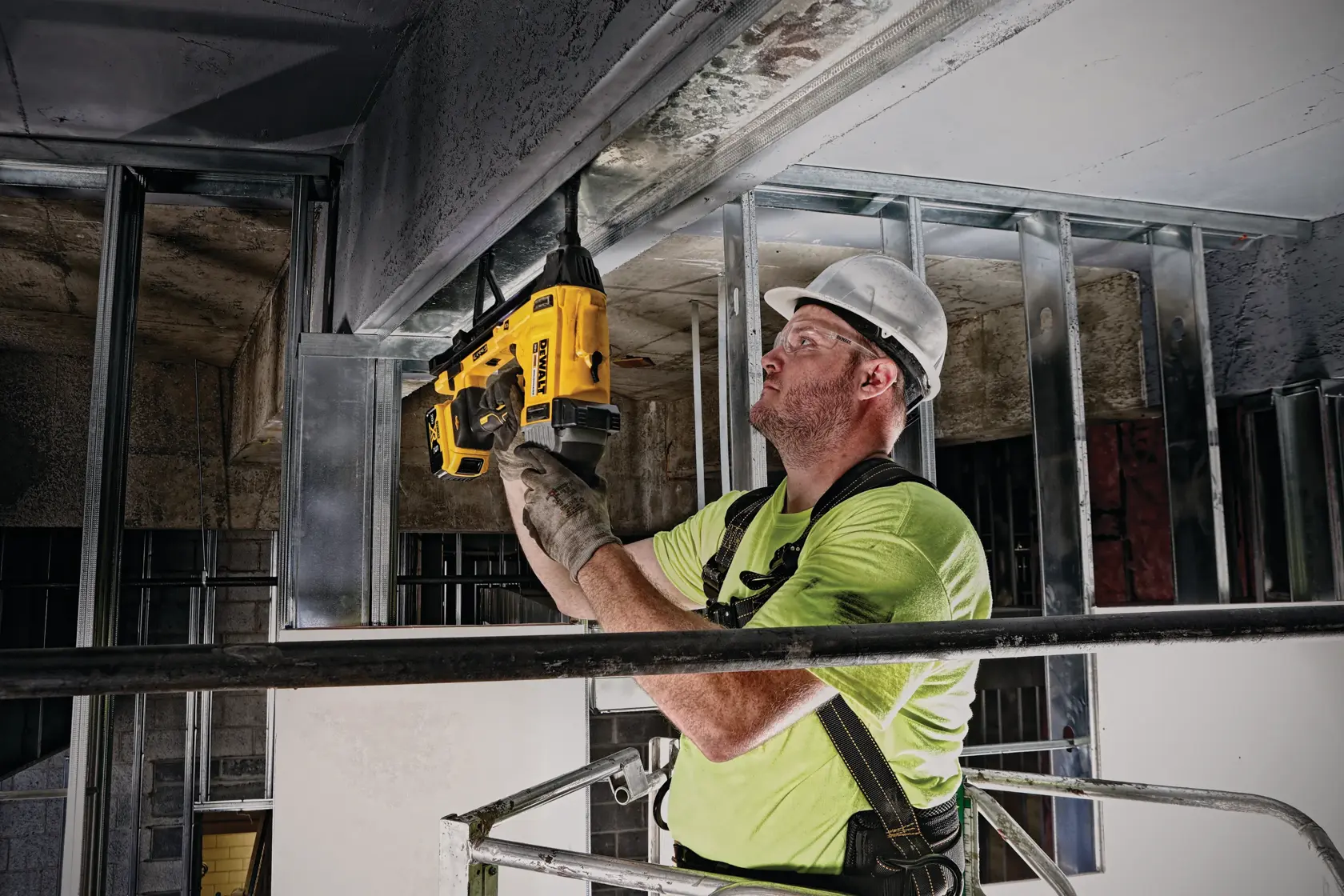 A worker wearing safety gear uses a DEWALT DCN891B cordless tool to fasten components on a metal ceiling framework at a construction site.