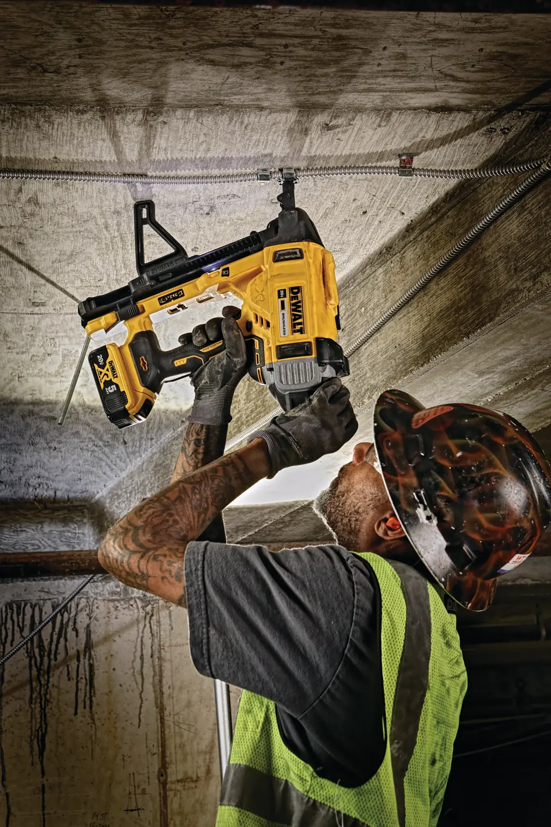 A construction worker wearing a safety vest and helmet uses a DEWALT DCN891B cordless tool to install wiring into a concrete ceiling.
