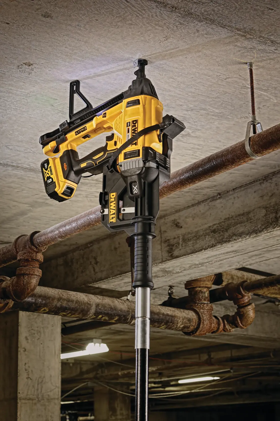 A DEWALT cordless tool, model DCN891B, attached to an extension pole and positioned against a concrete ceiling for fastening, likely in an industrial or parking garage setting. The tool is yellow and black and is designed for overhead applications.
