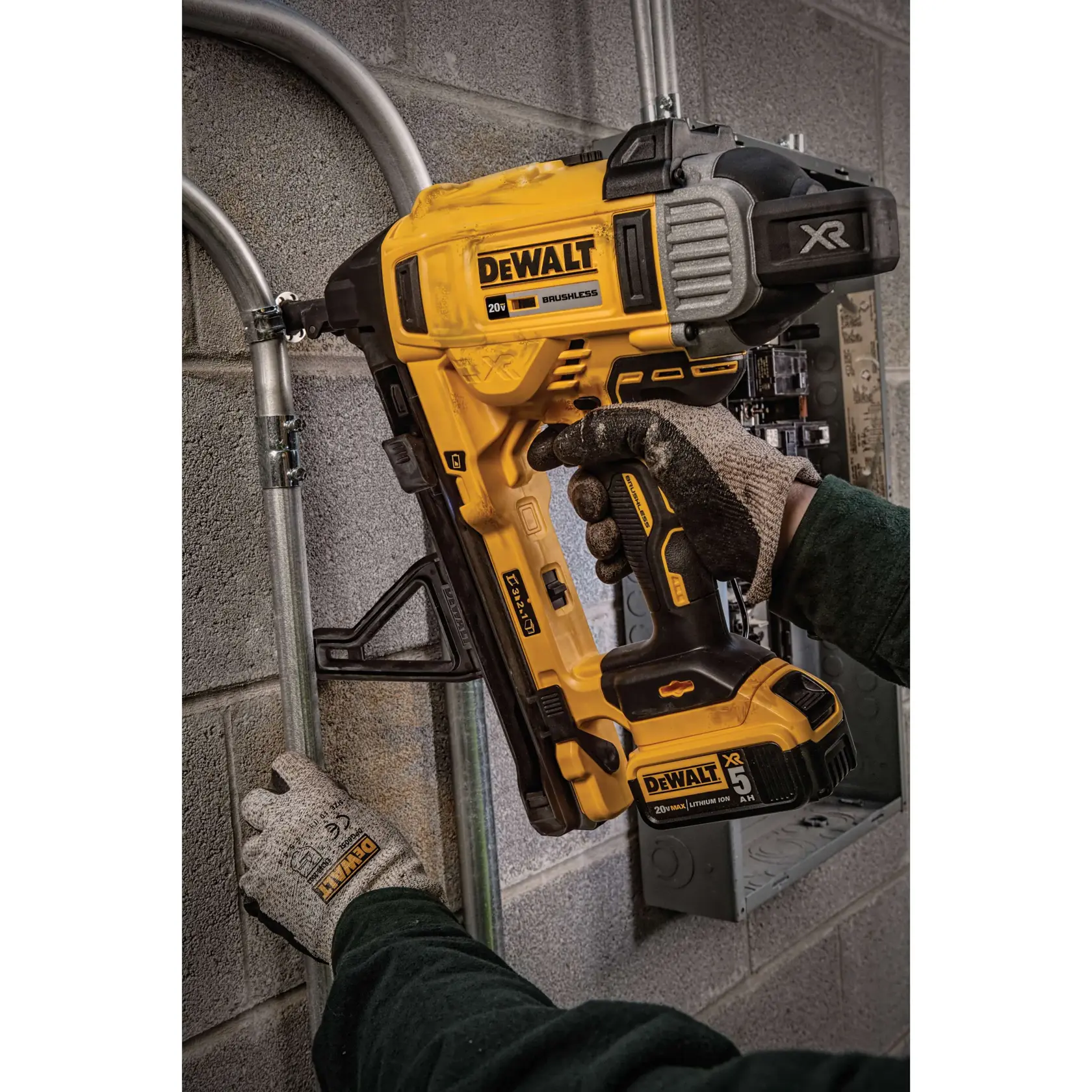 A person wearing work gloves uses a DEWALT DCN891P2 20V cordless tool to secure metal pipes to a concrete wall. The tool is yellow and black, with labels indicating it is brushless and part of the XR series.