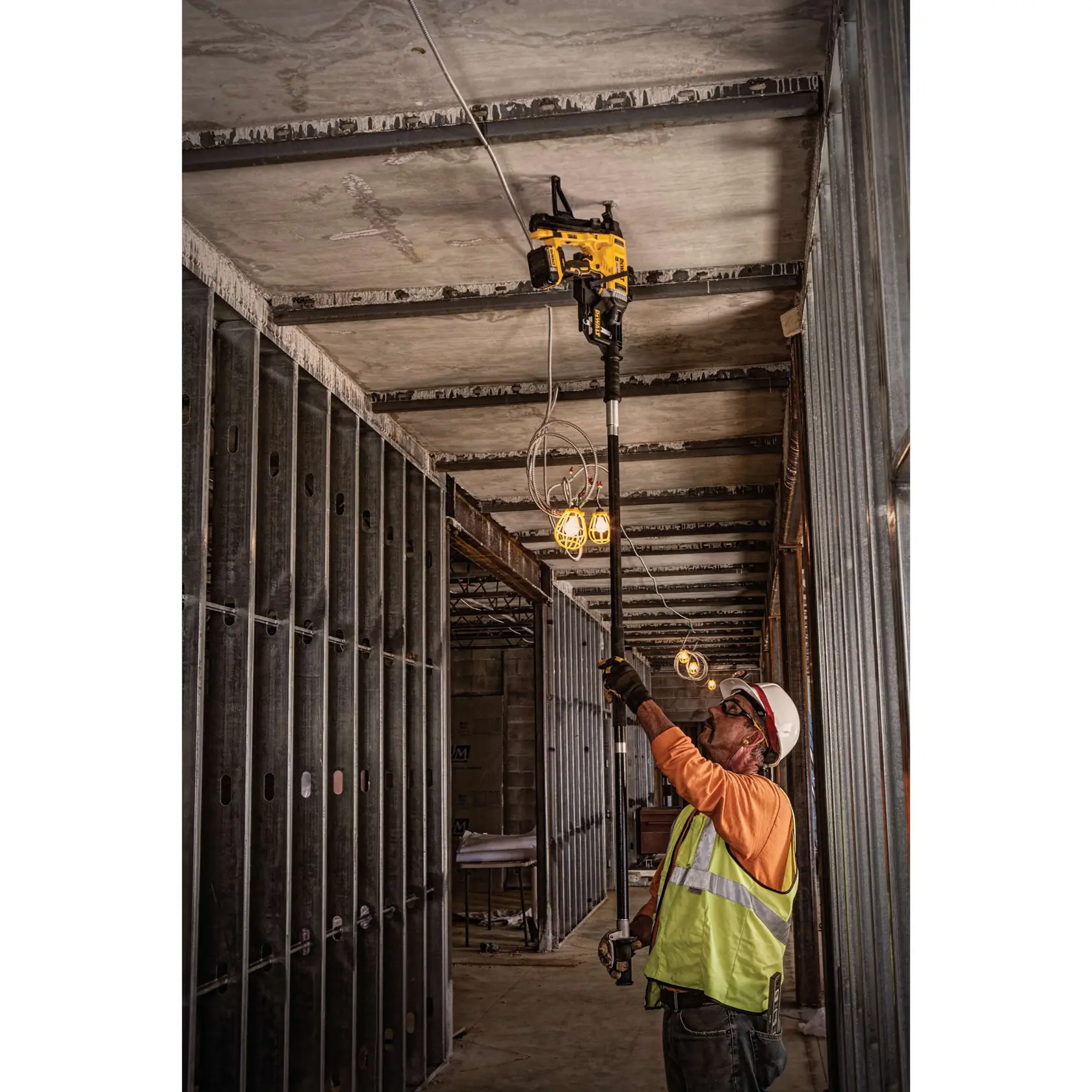 A construction worker wearing a safety vest and hard hat uses a DEWALT cordless tool to fasten conduit to the ceiling in an unfinished building with exposed metal framing and overhead lights.