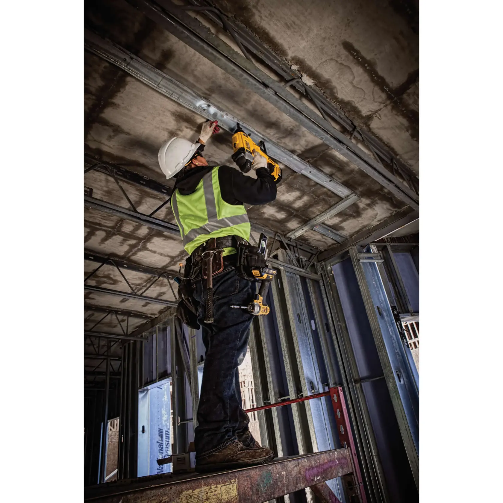 A construction worker wearing a safety vest and hard hat stands on a platform and uses a DEWALT power tool to secure metal framing to a concrete ceiling in an unfinished building.