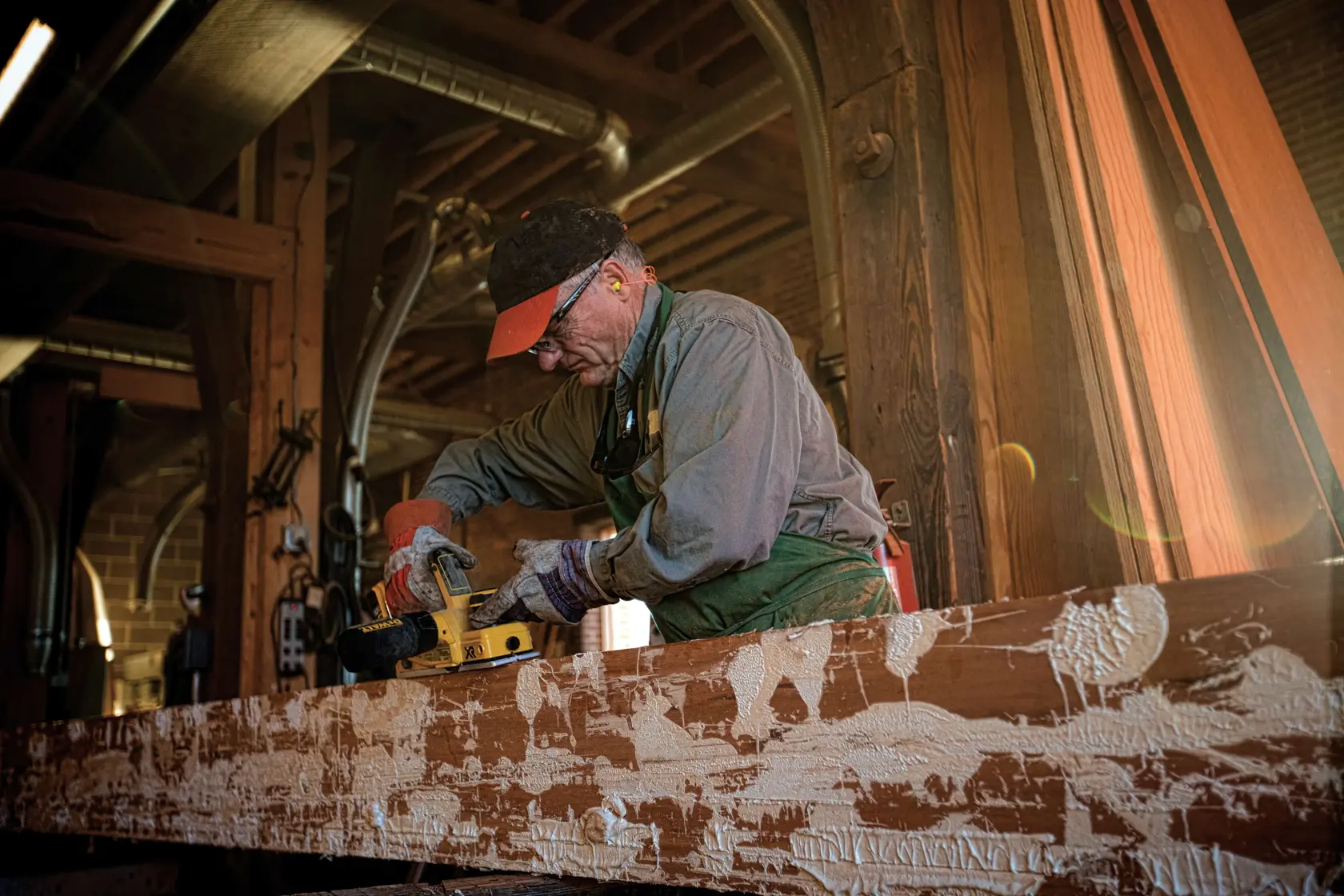 A person using a DEWALT DCP580B cordless planer on a wooden beam in a workshop. The individual is wearing gloves and work attire. The background shows workshop equipment and wooden materials.