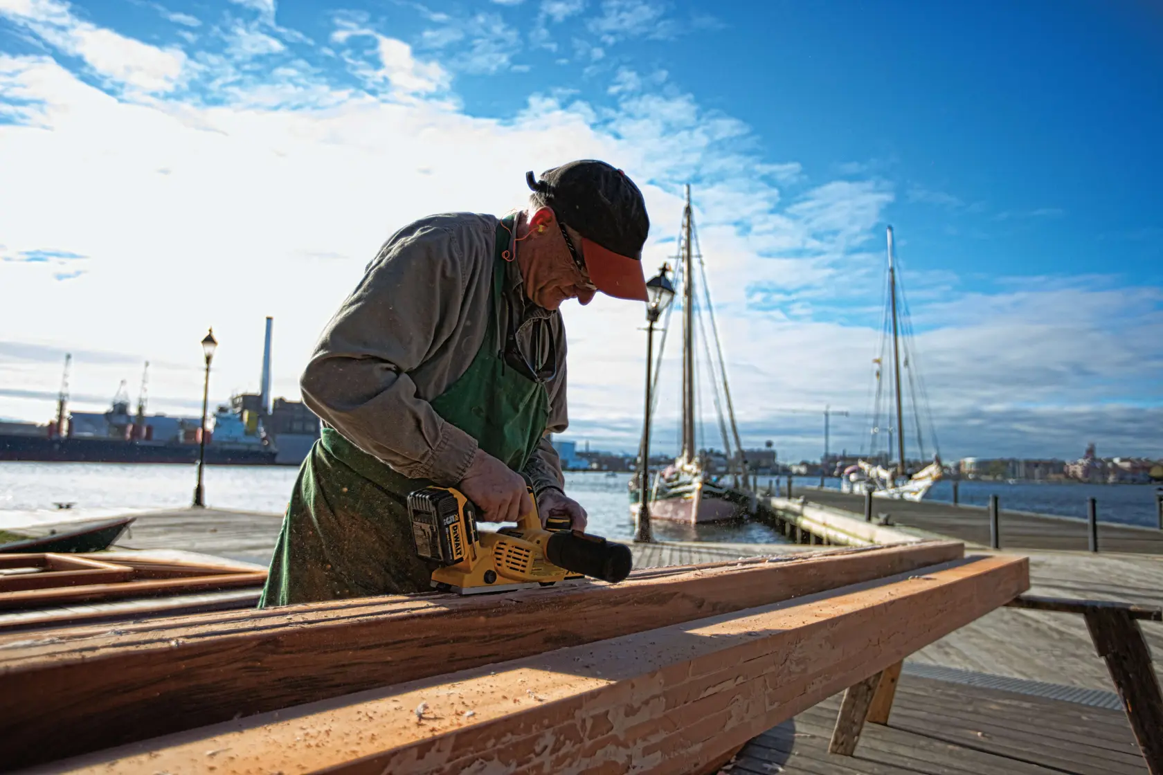 A person is using a DEWALT DCP580B power tool to plane a wooden beam outdoors near a waterfront with boats and industrial buildings in the background. The individual's face is blurred.