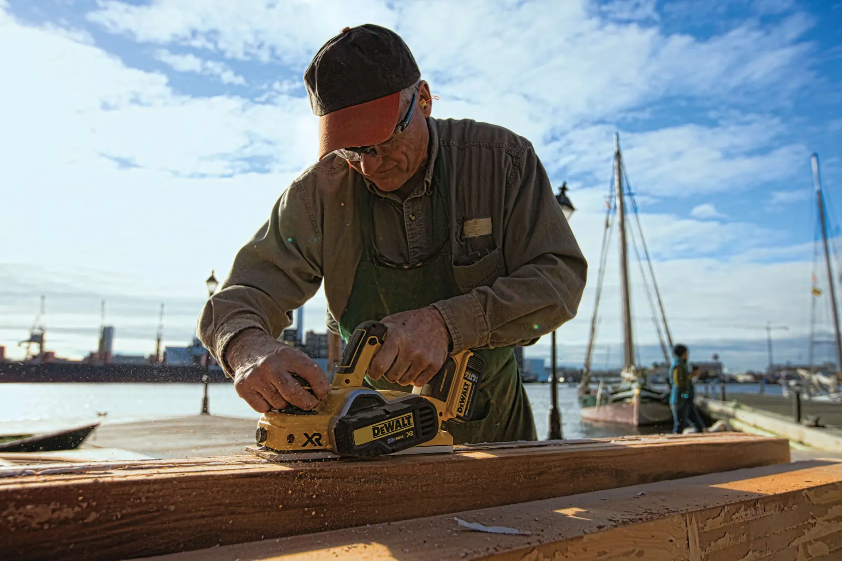 A person is using a DEWALT DCP580B cordless planer to smooth a piece of wood outdoors near a marina, with boats and water visible in the background. The person's face is obscured.