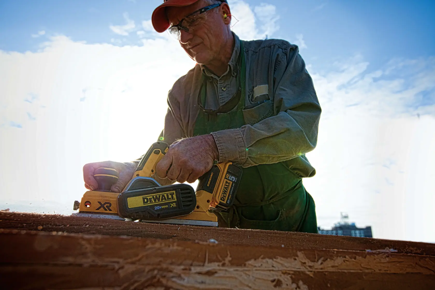 A person using a DEWALT DCP580B cordless planer, working on a piece of wood outdoors under a partly cloudy sky. The person's face is blurred for privacy.