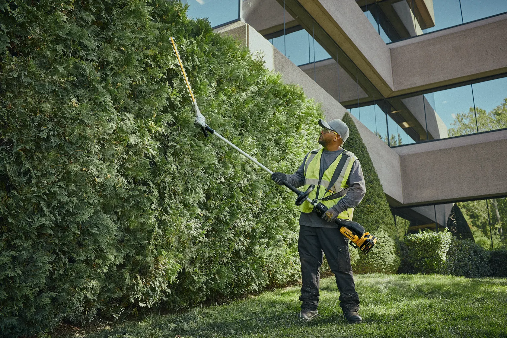 A person wearing safety gear is using a DEWALT pole hedge trimmer to trim tall bushes in front of a modern building.