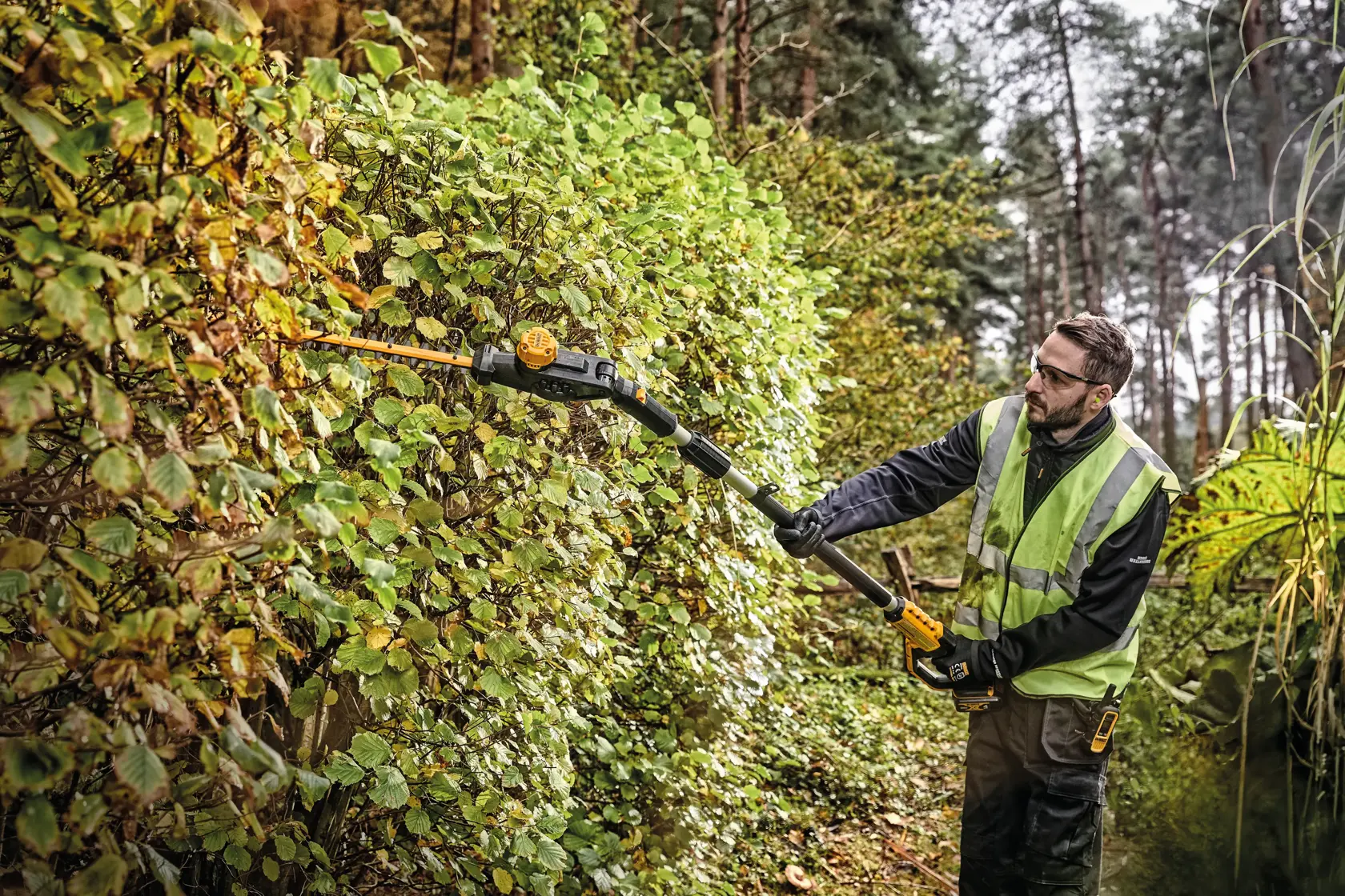 A person wearing a reflective safety vest is using a DEWALT DCPH820B pole hedge trimmer to cut tall hedges outdoors in a wooded area. Their face is blurred for privacy.