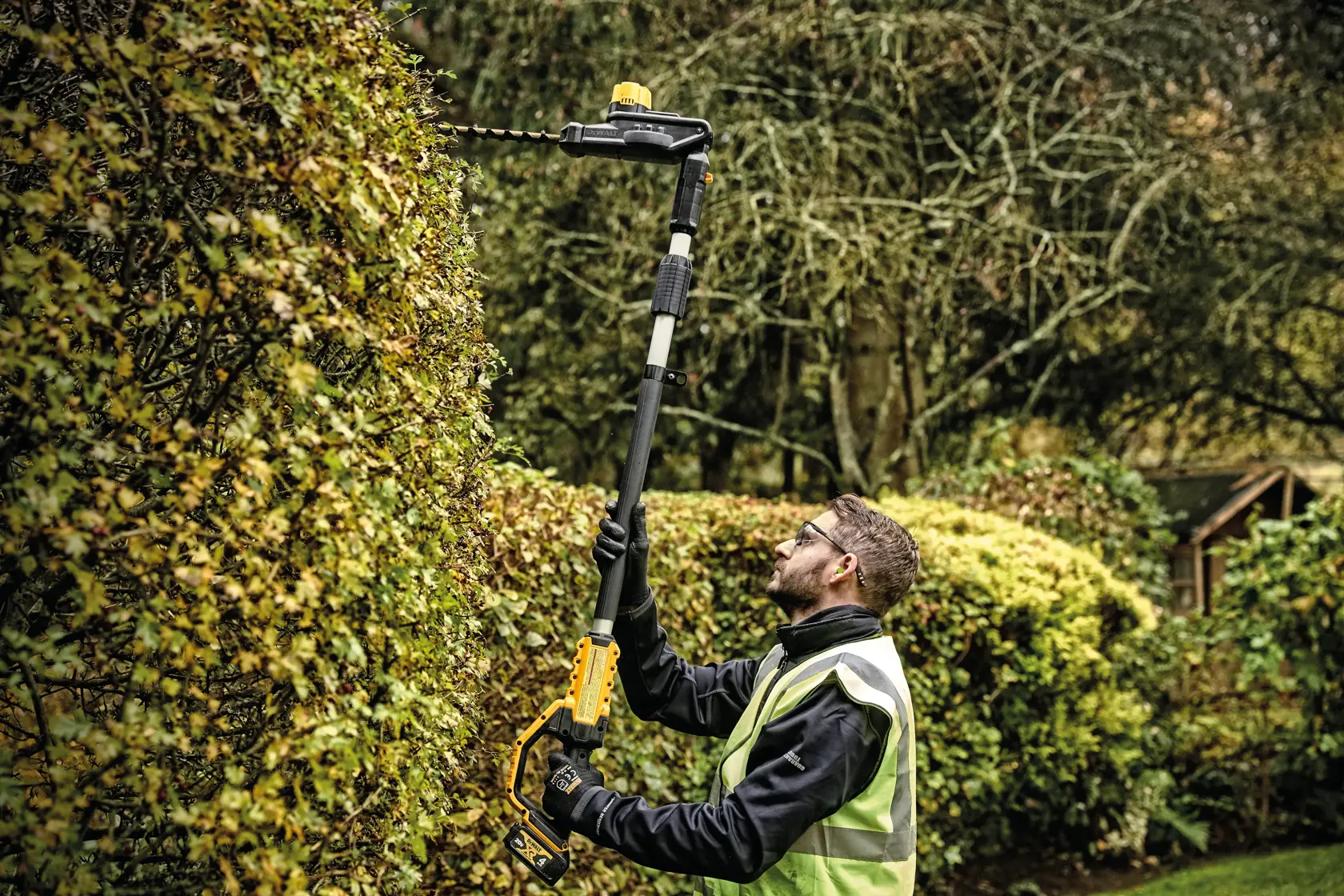A person using a DEWALT DCPH820B cordless pole hedge trimmer to trim tall hedges in a garden. The tool has a long, extendable pole for reaching high branches, and the person is wearing protective gloves and a safety vest.