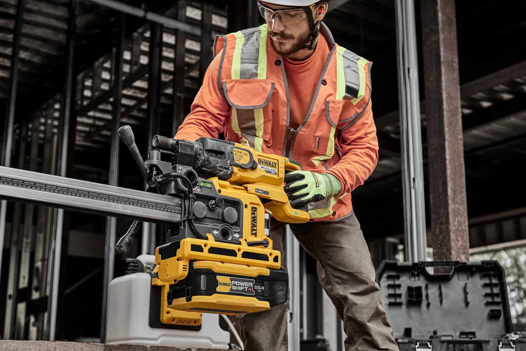 A construction worker wearing an orange safety jacket and gloves operates a yellow DEWALT power tool, specifically a heavy-duty drill or driver, inside a building under construction. The tool is being used on a metal beam, and a black tool case is visible nearby.